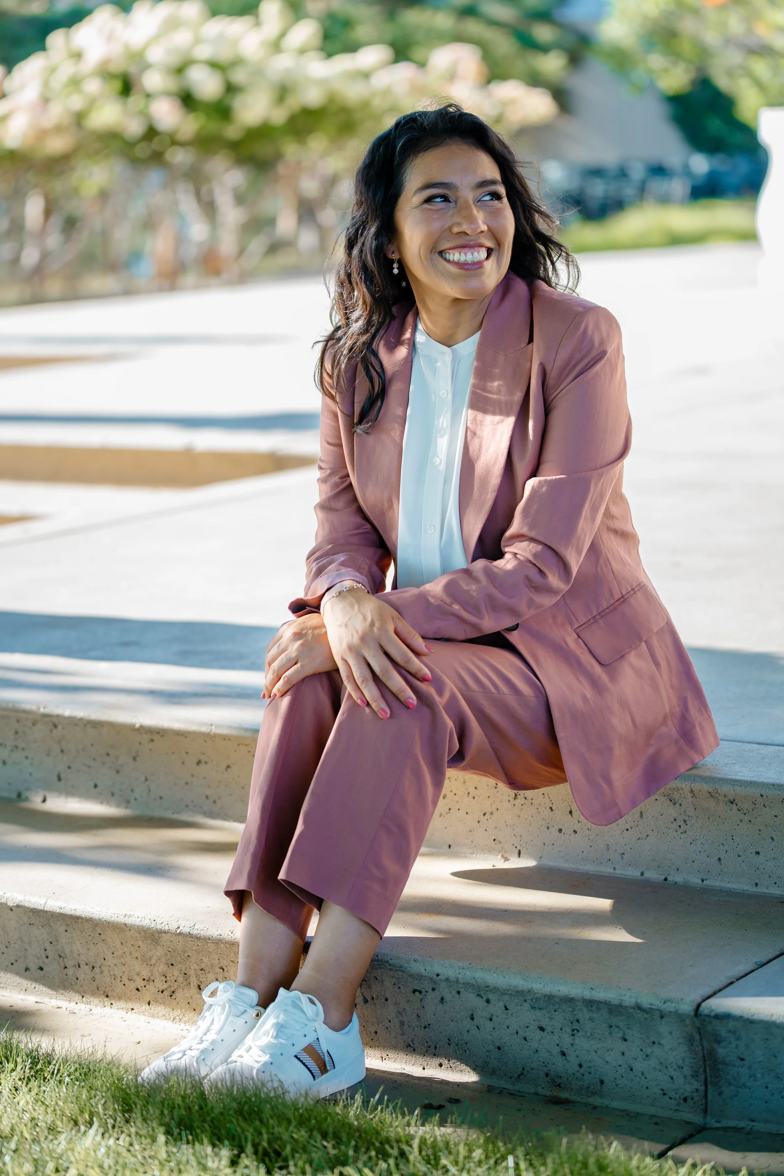 A woman sitting on a concrete ledge outdoors, smiling and looking to her left. She is wearing a pink suit with white sneakers and has dark curly hair.