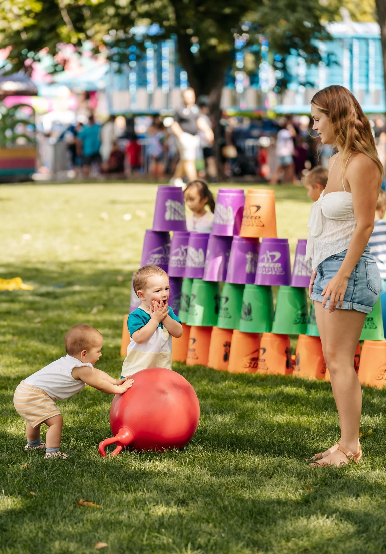 Children playing with a large red ball at a carnival or fair, with a woman supervising, and colorful cups stacked in the background.