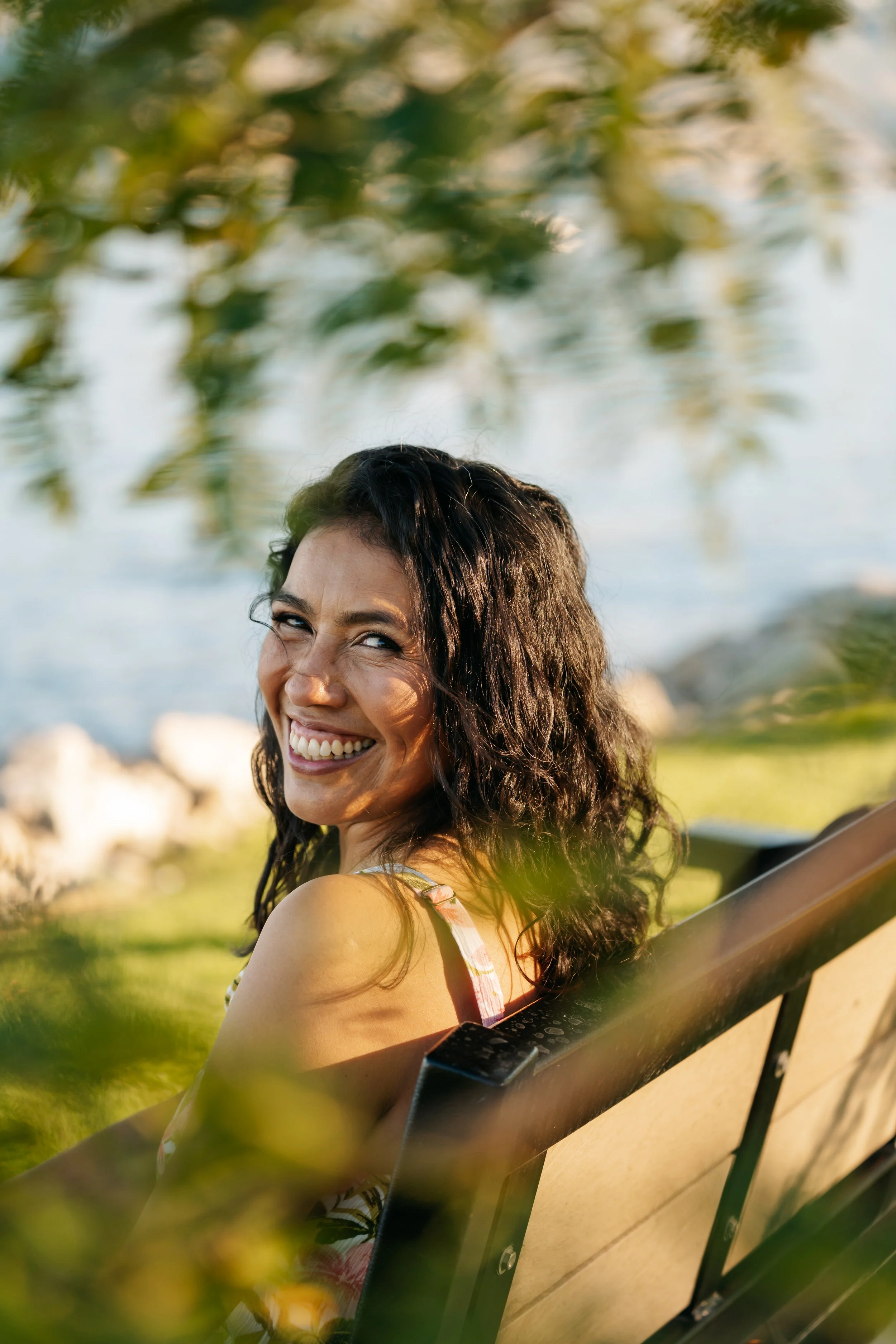 Smiling woman sitting on park bench near water, with trees and a sunset or sunrise in the background.