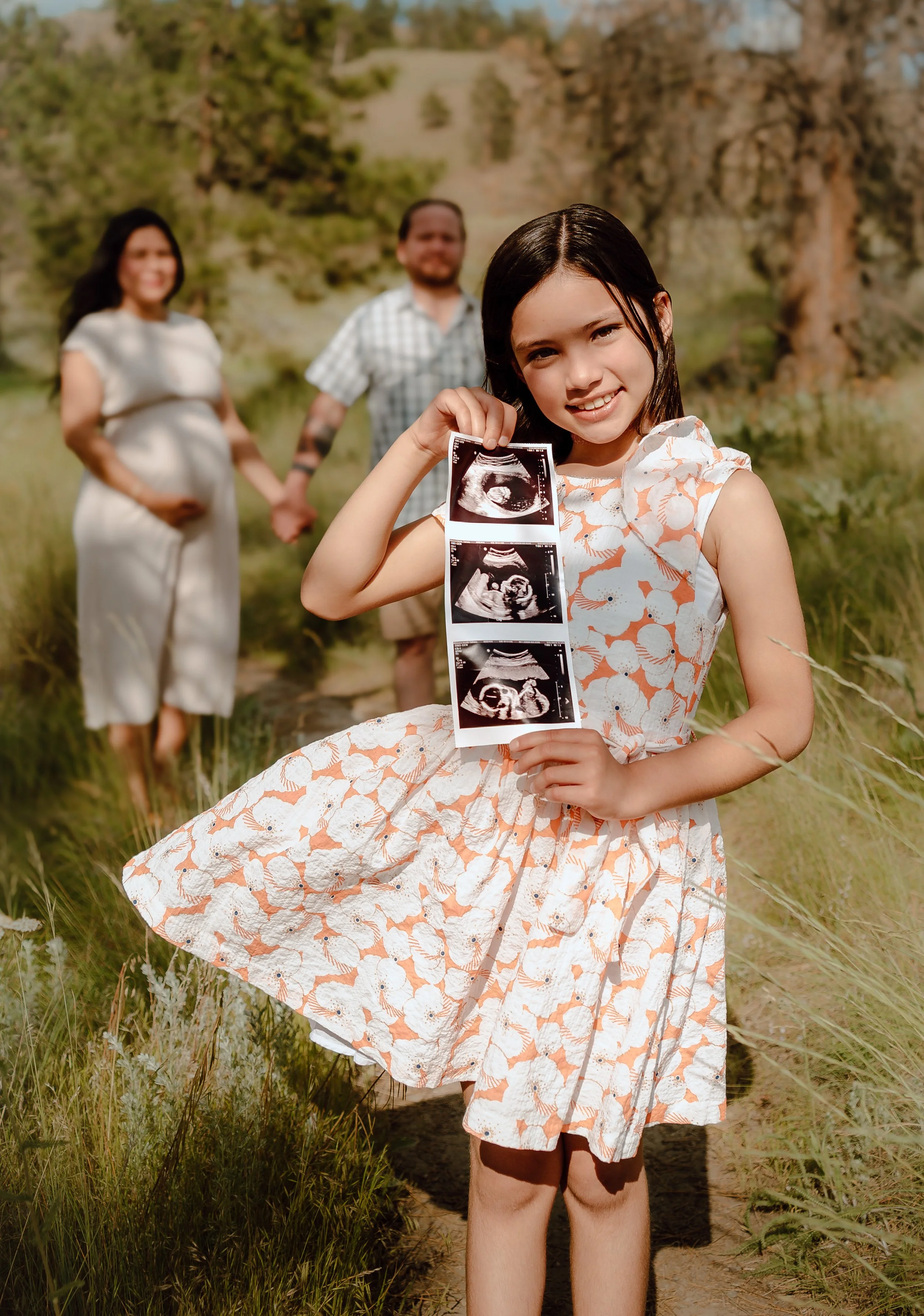 A young girl holding ultrasound images, smiling, with her pregnant mother and father in the background in an outdoor natural setting.