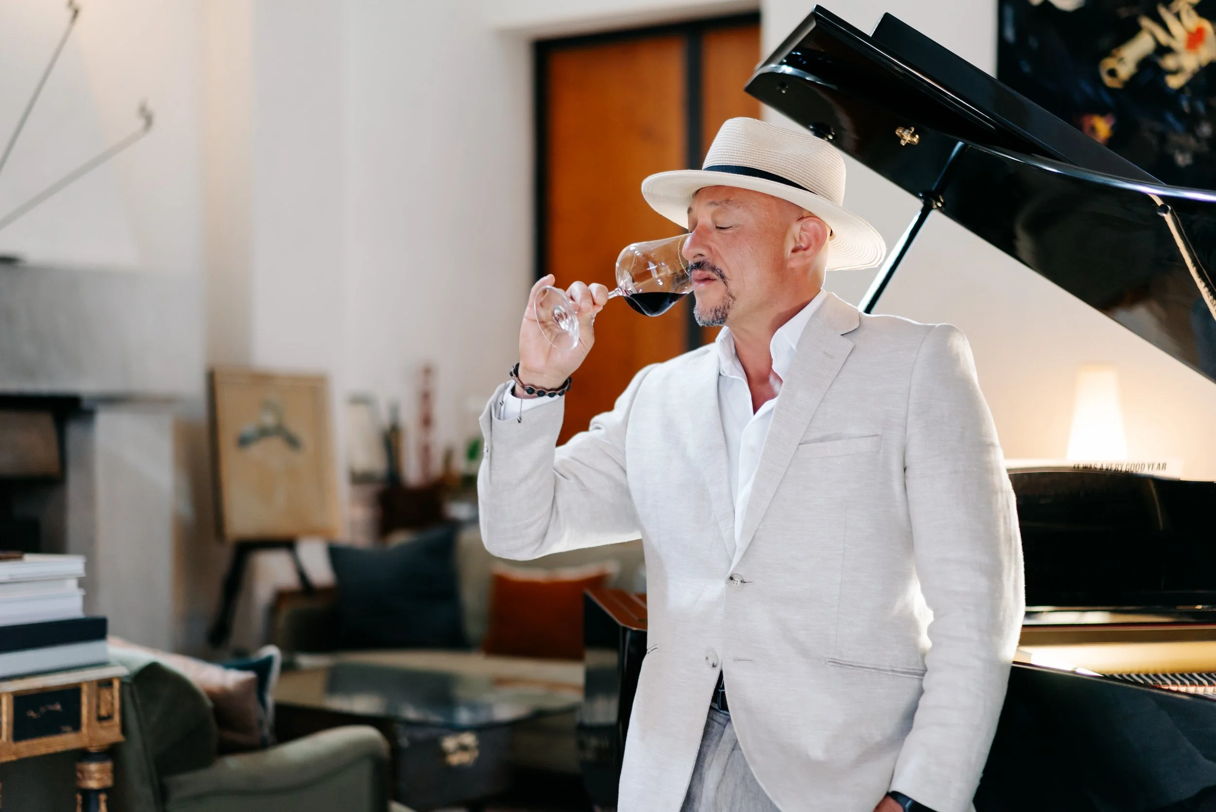 A man in a beige suit and white shirt wearing a wide-brimmed hat, drinking red wine near a black grand piano in a cozy living room.
