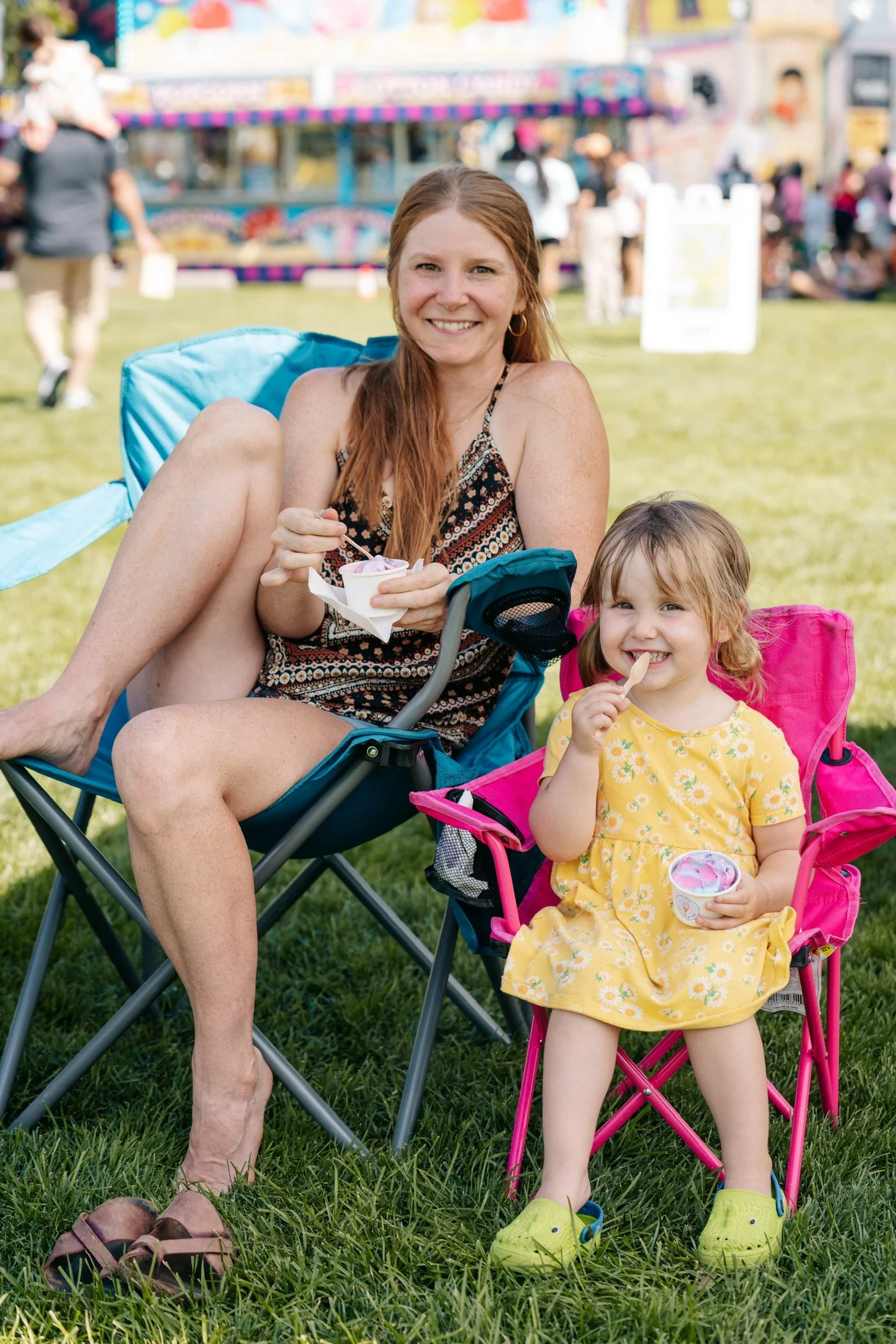 A woman and a young girl sitting on colorful camping chairs outdoors, enjoying ice cream at a fair or festival, with a carnival ride visible in the background.