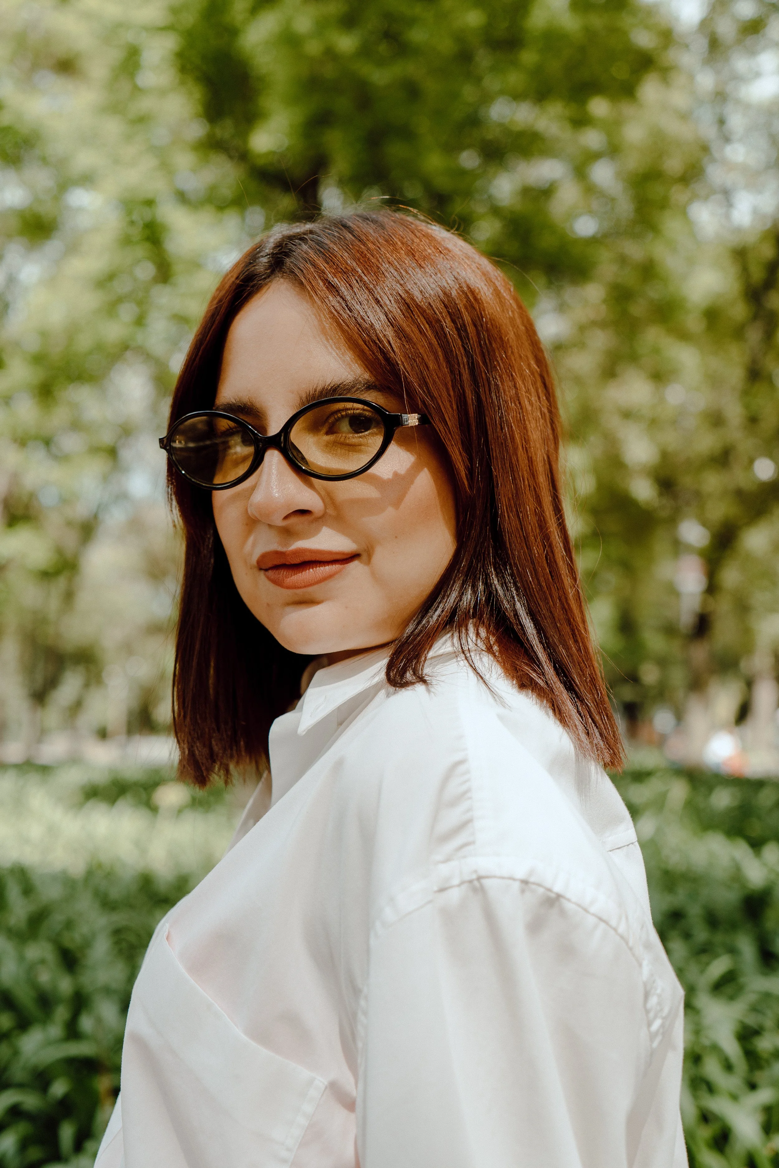 A woman with shoulder-length dark brown hair and glasses, wearing a white shirt, standing outdoors with greenery and trees in the background.