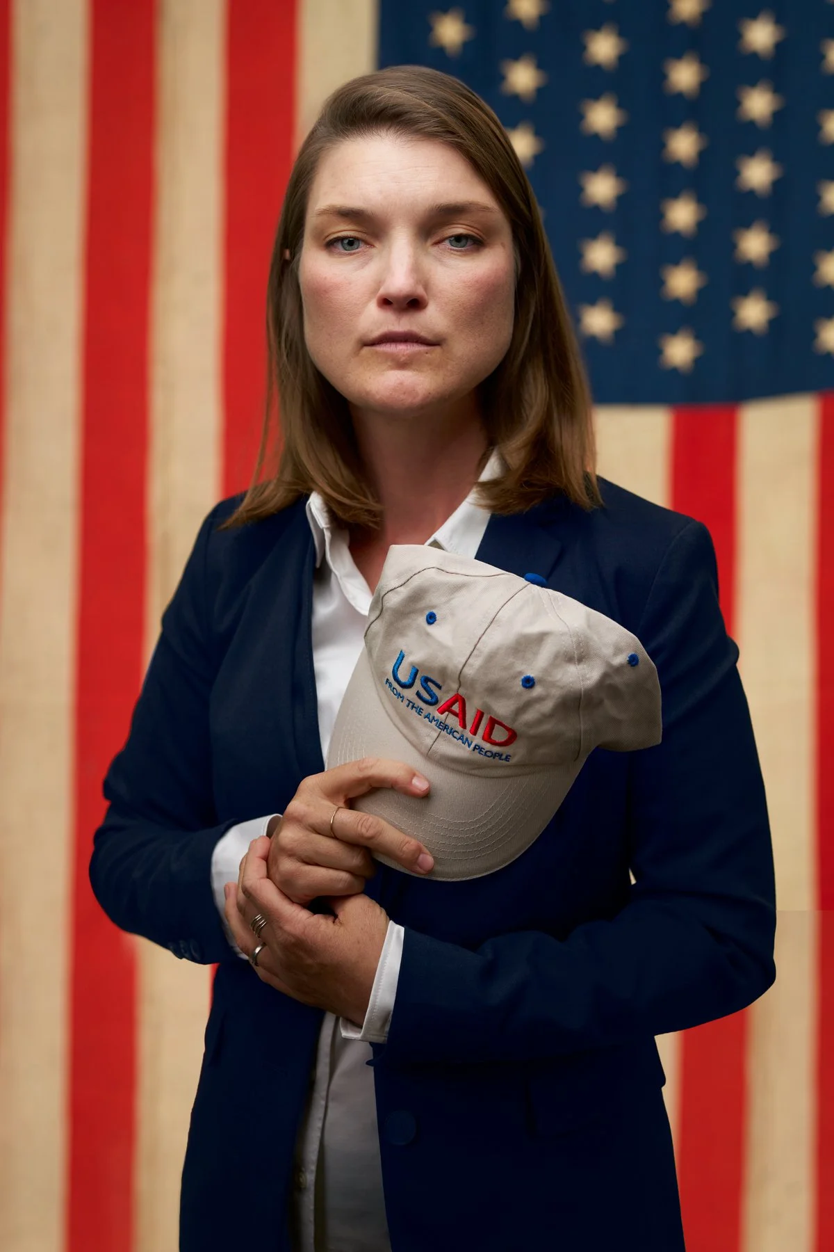 A woman in a navy blazer and white shirt holding a beige baseball cap with the text 'USAID from the American People' embroidered on it. Behind her is a large American flag.