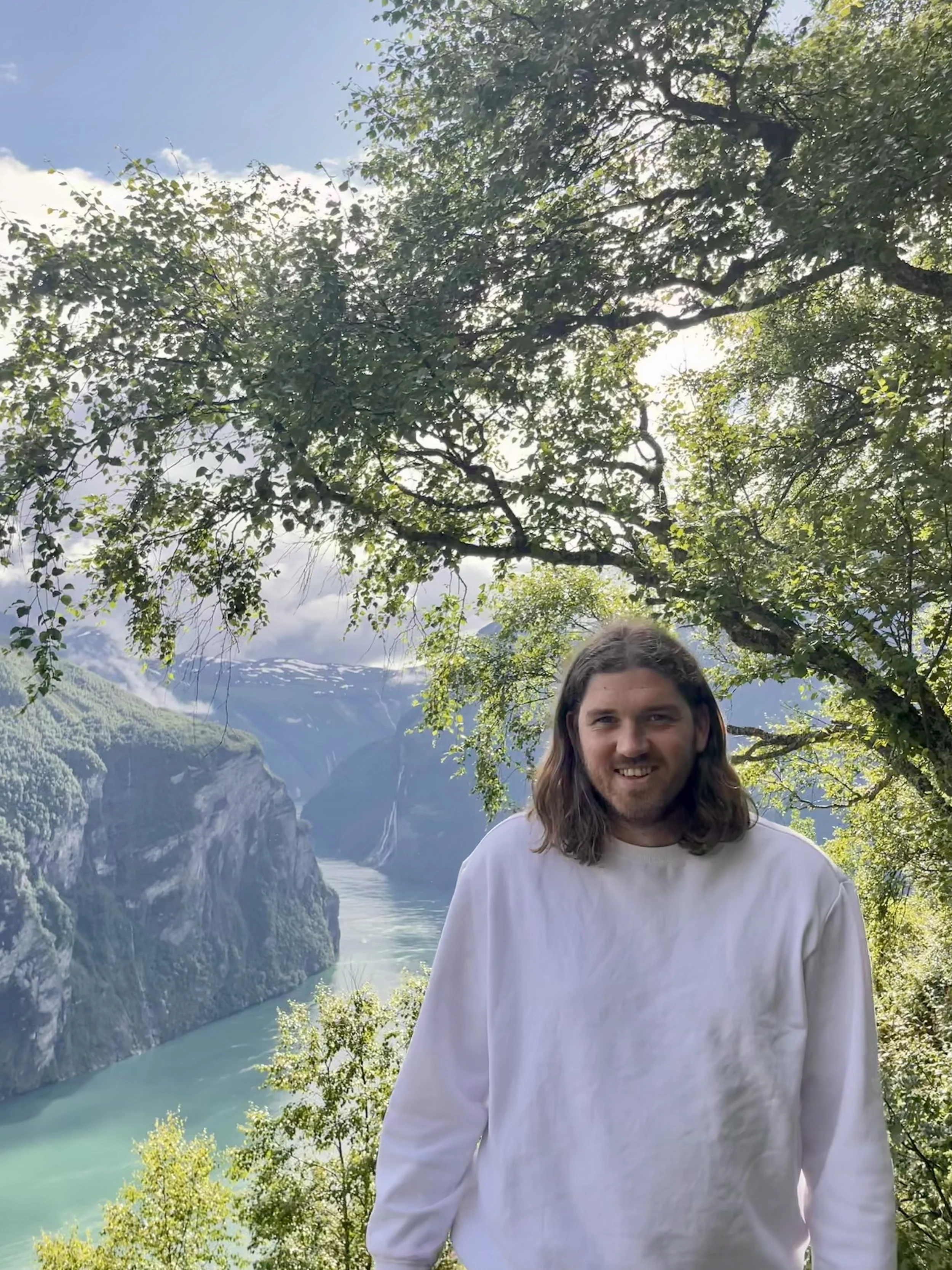 En man med långt hår och vit tröja står framför en naturskön fjord med höga berg och klart vatten, under en grön trädgren i ett fjärran landskap.