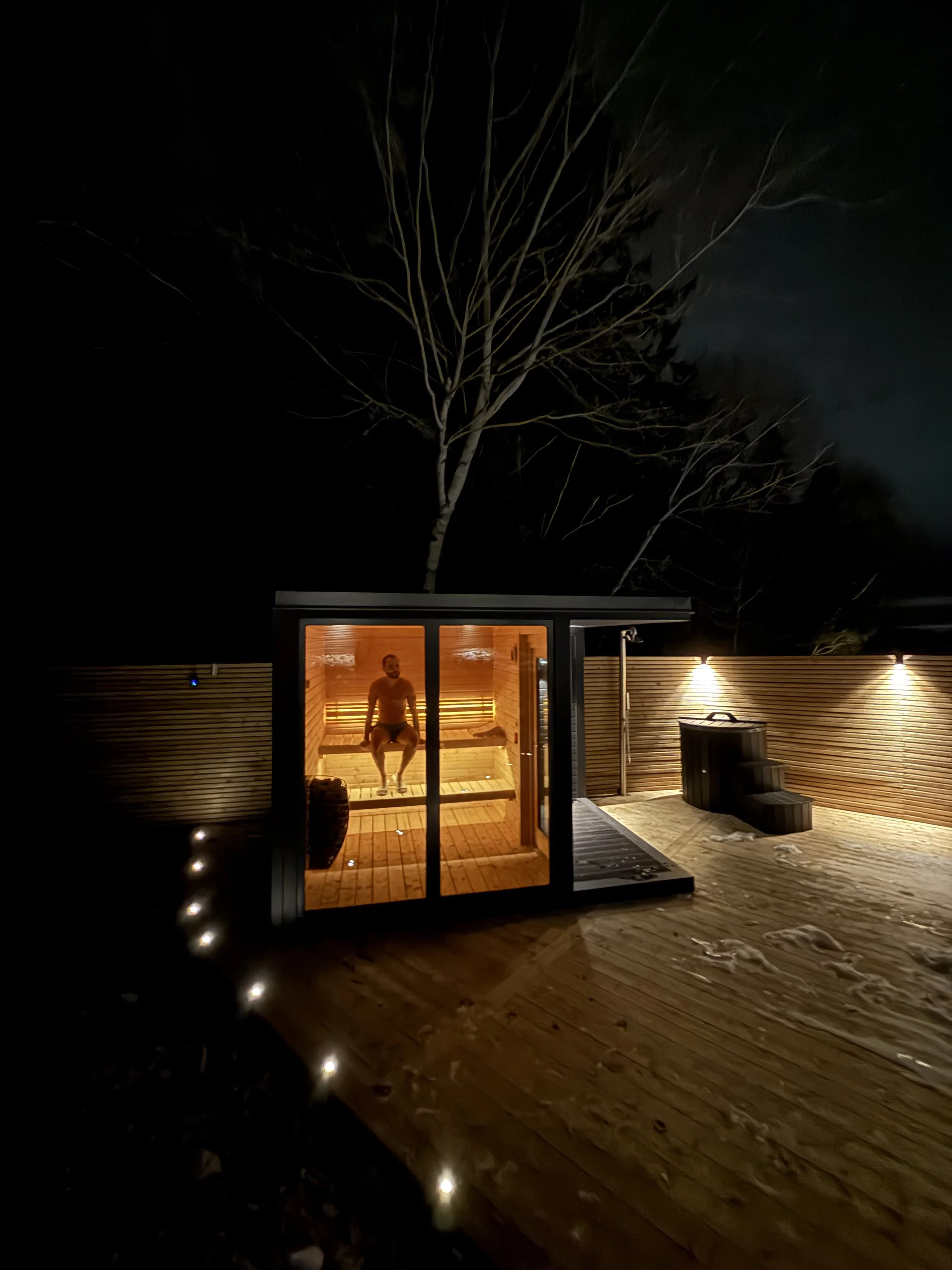 En person sitter i ett infrarött badhus på kvällen, omgivet av en träaltan och en trävägg, med belysning i mörkret och en stor träd i bakgrunden.