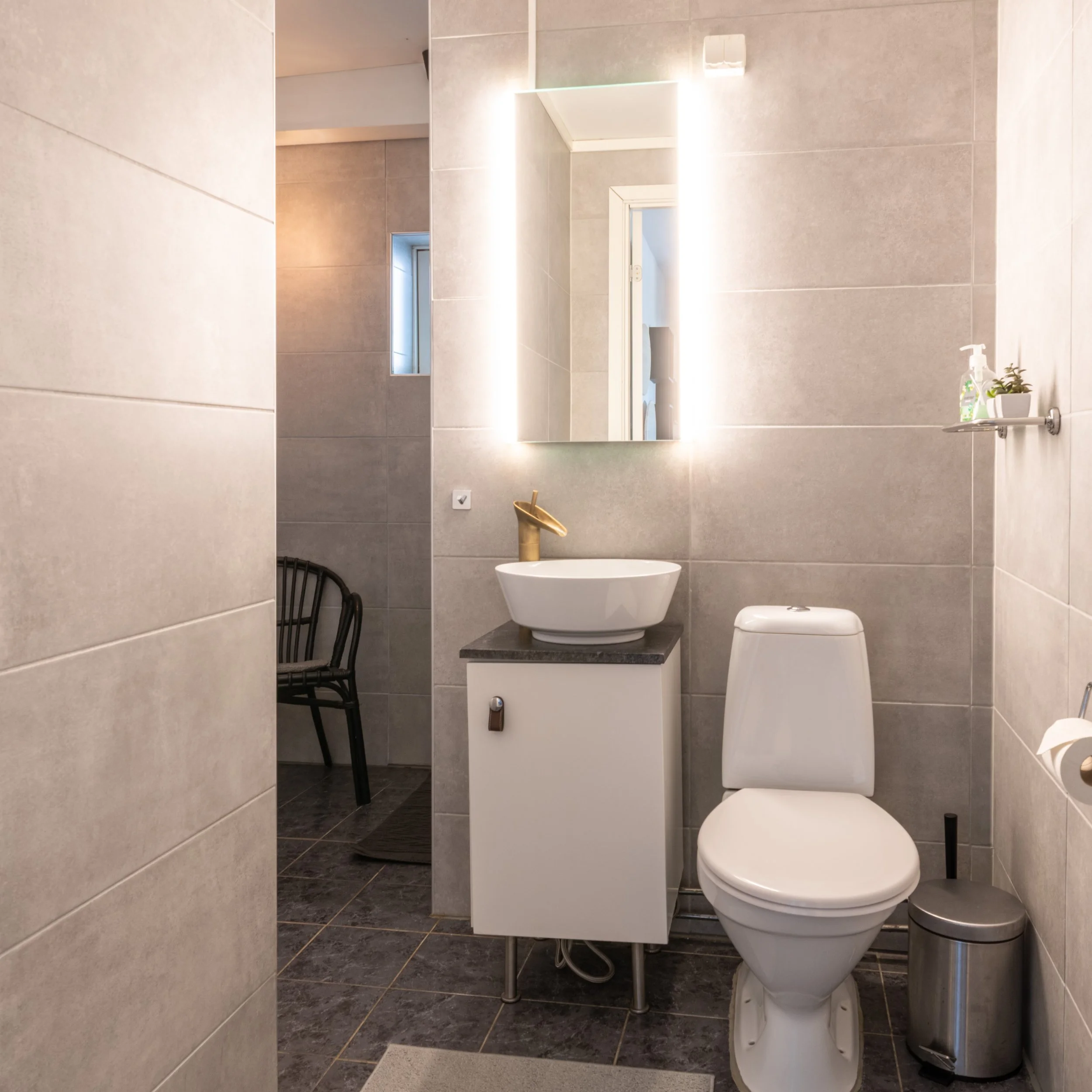 En modern toalett med spegel med lysdioder, handfat med guldarmatur, toalett, spegel, liten växt, tvål, pappersrulle och sopkorg.