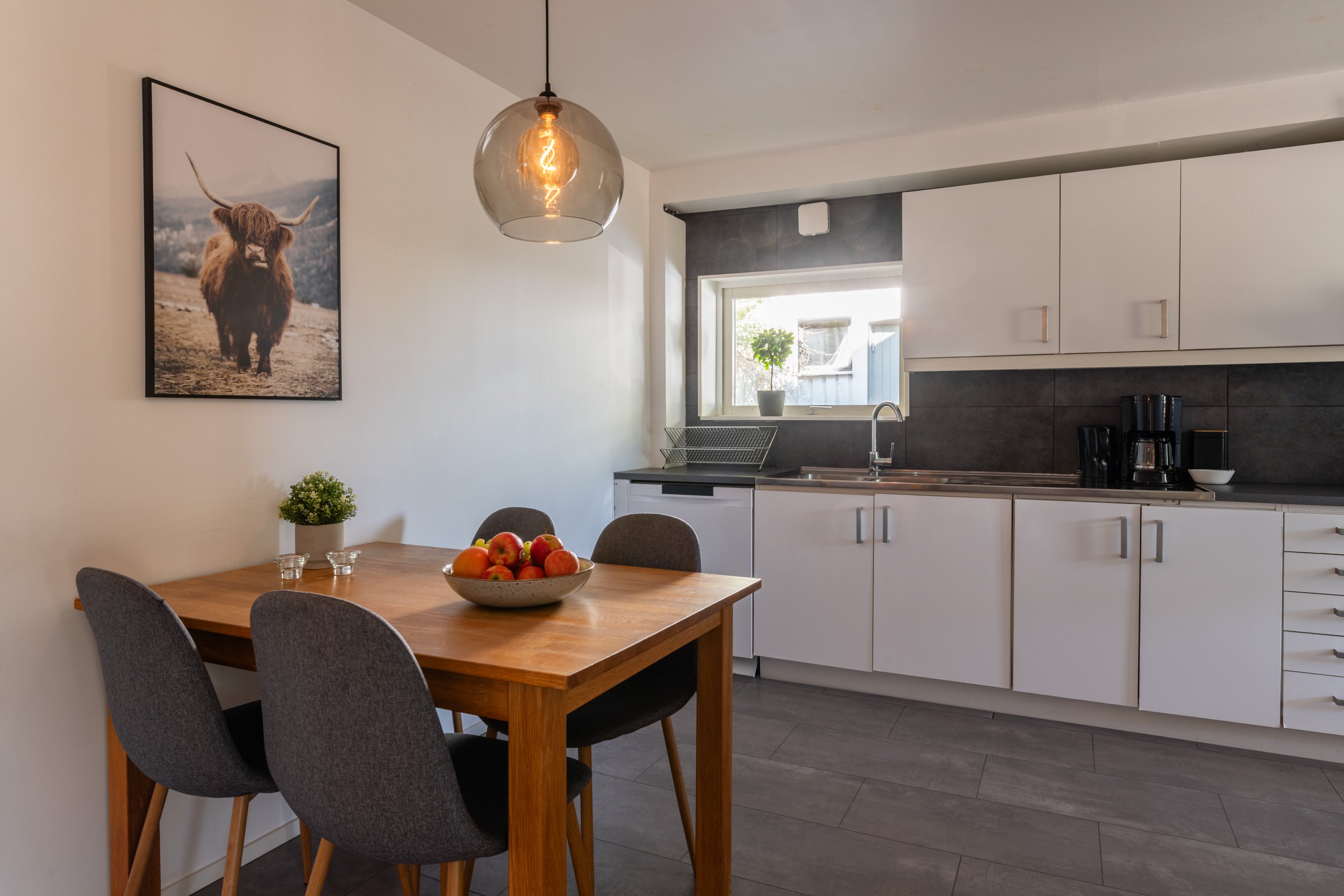 Modern köksdel med ett matbord, fyra stolar, ett fönster, växt och plans myt på vägg.