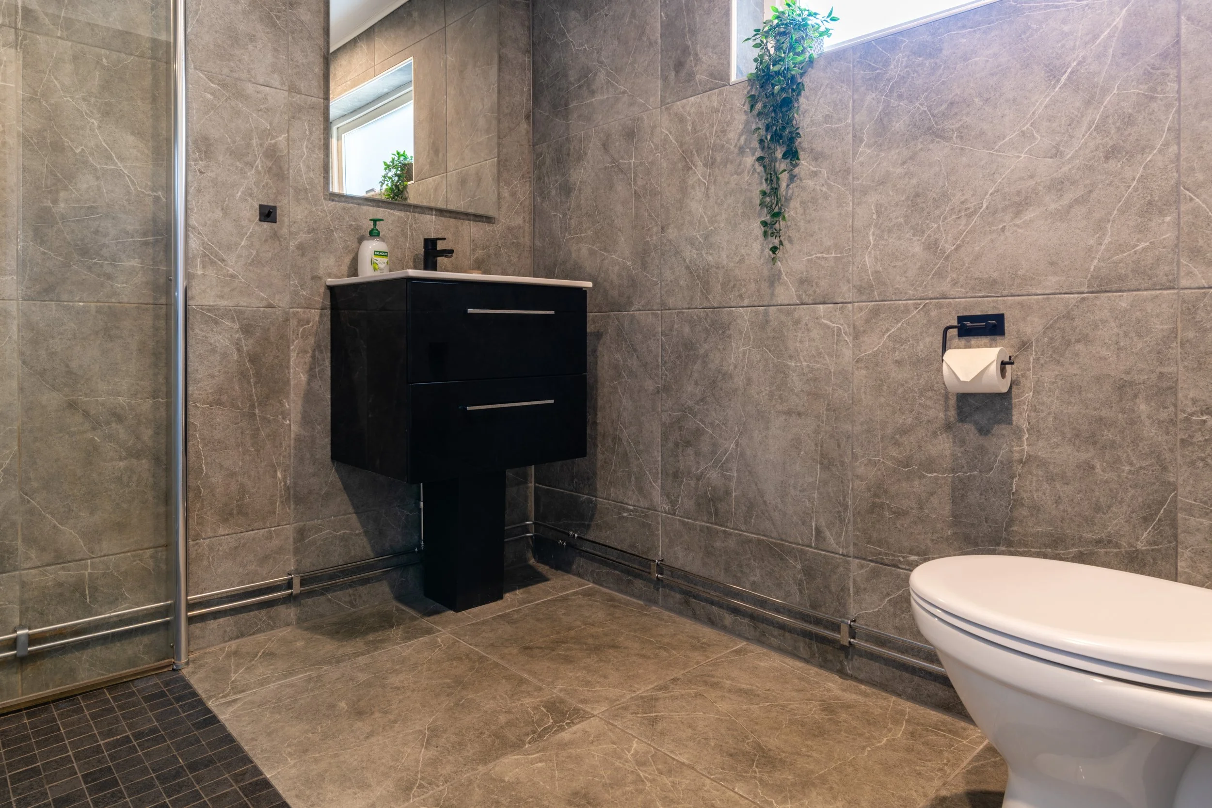 Badrum med grå kakelväggar och golv, svart handfat med spegel, fönster med gröna växter, toalett med toarulle, växt hängande på vägg