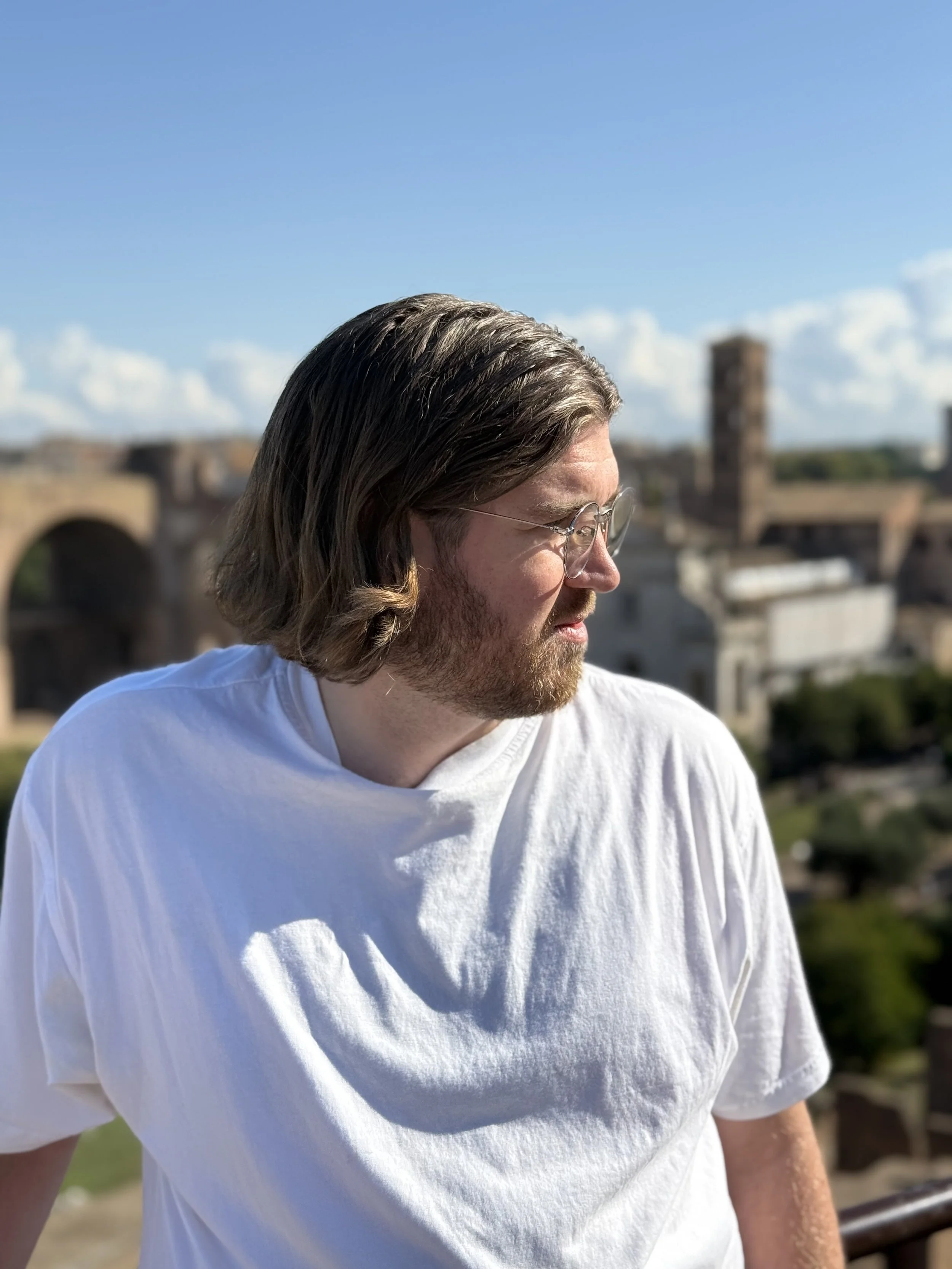 Man med långt brunt hår och glasögon tittar åt höger, med romerska ruiner i bakgrunden under en klar blå himmel.