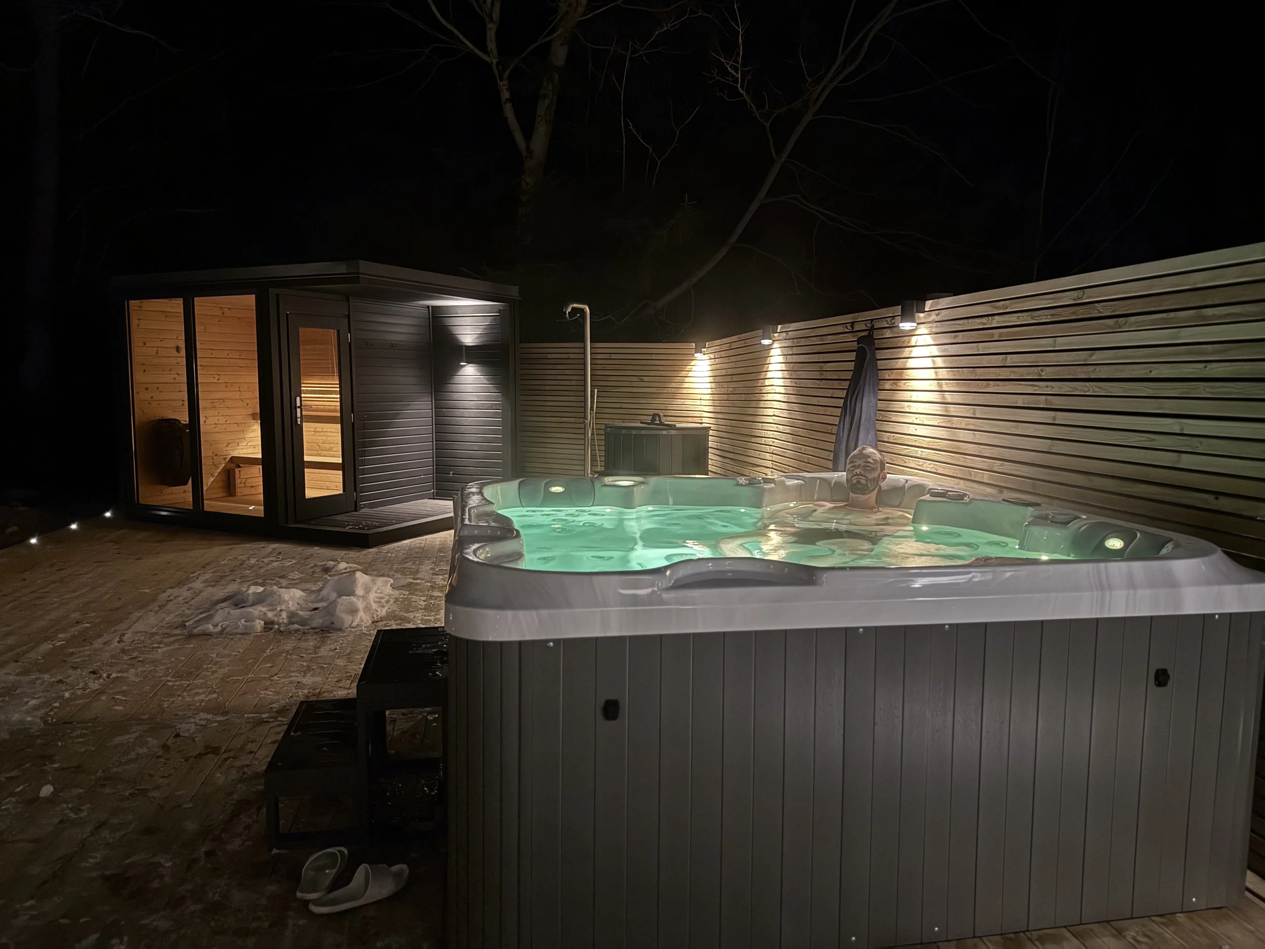 Man som sitter i ett utomhusiskt jacuzzi på natten, omgivet av en träräckt vägg med utomhusbelysning och en bastubyggnad i bakgrunden.