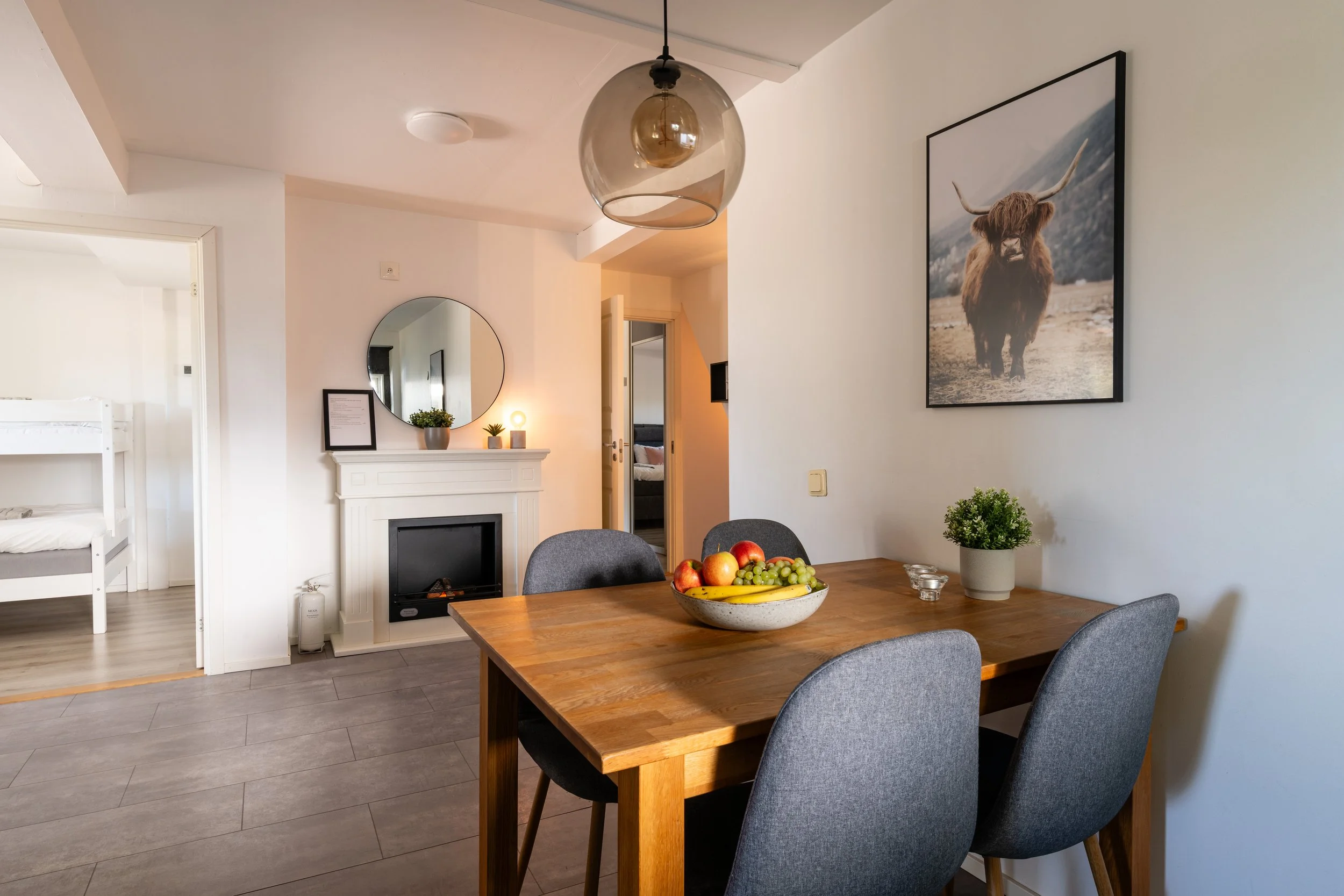 Matsal med träbord, fyra grå stolar, fruktfat, växt och tavla med yakt i form av en bäver, eldstad i bakgrunden, ljus och modernt inrett.