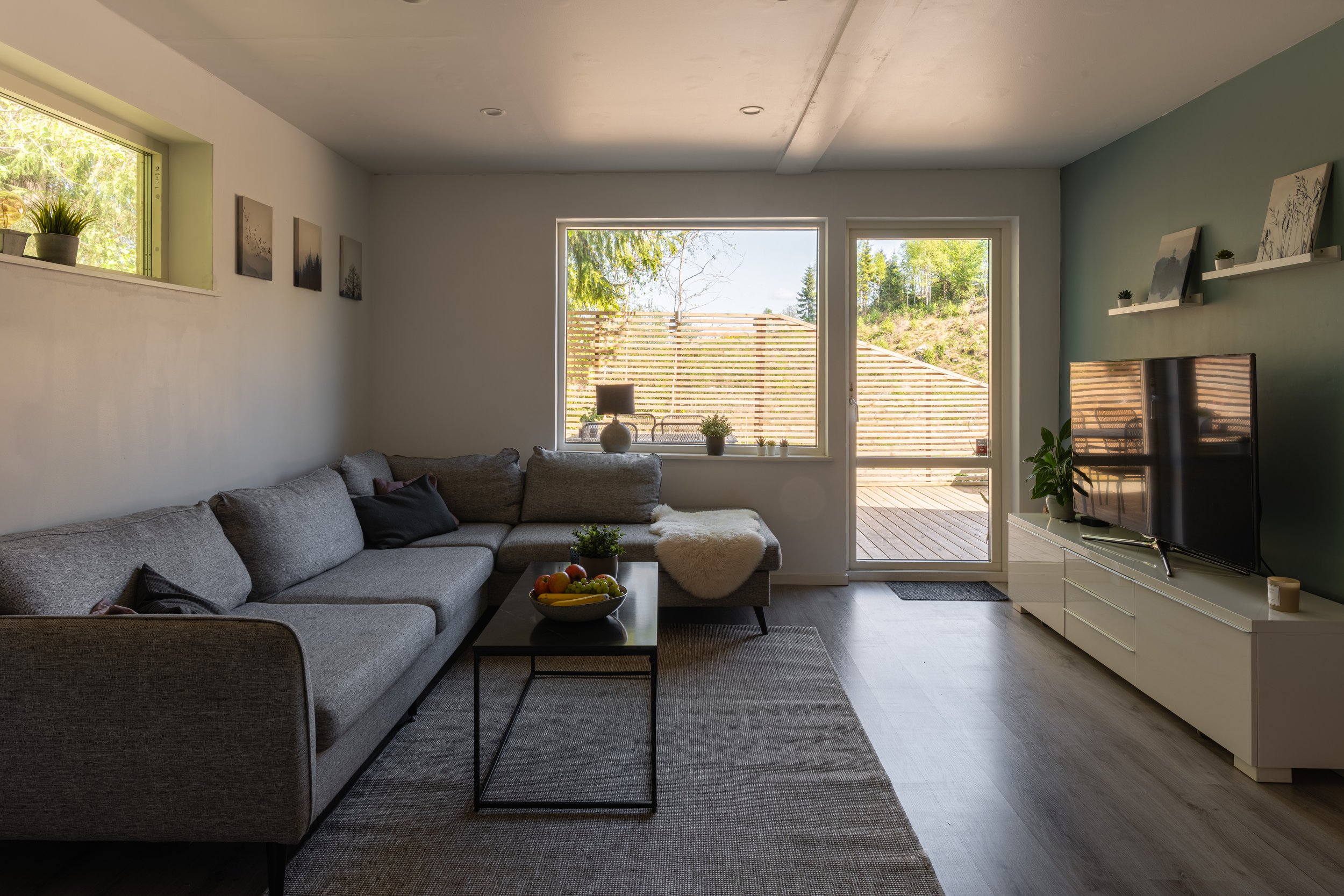 Ljust vardagsrum med stor grå hörnsoffa, filt, svart soffkuddar, ett svart soffbord med fruktskål, tv på vit mediabänk, växter, och fönster med utsikt mot träd och uteplats.