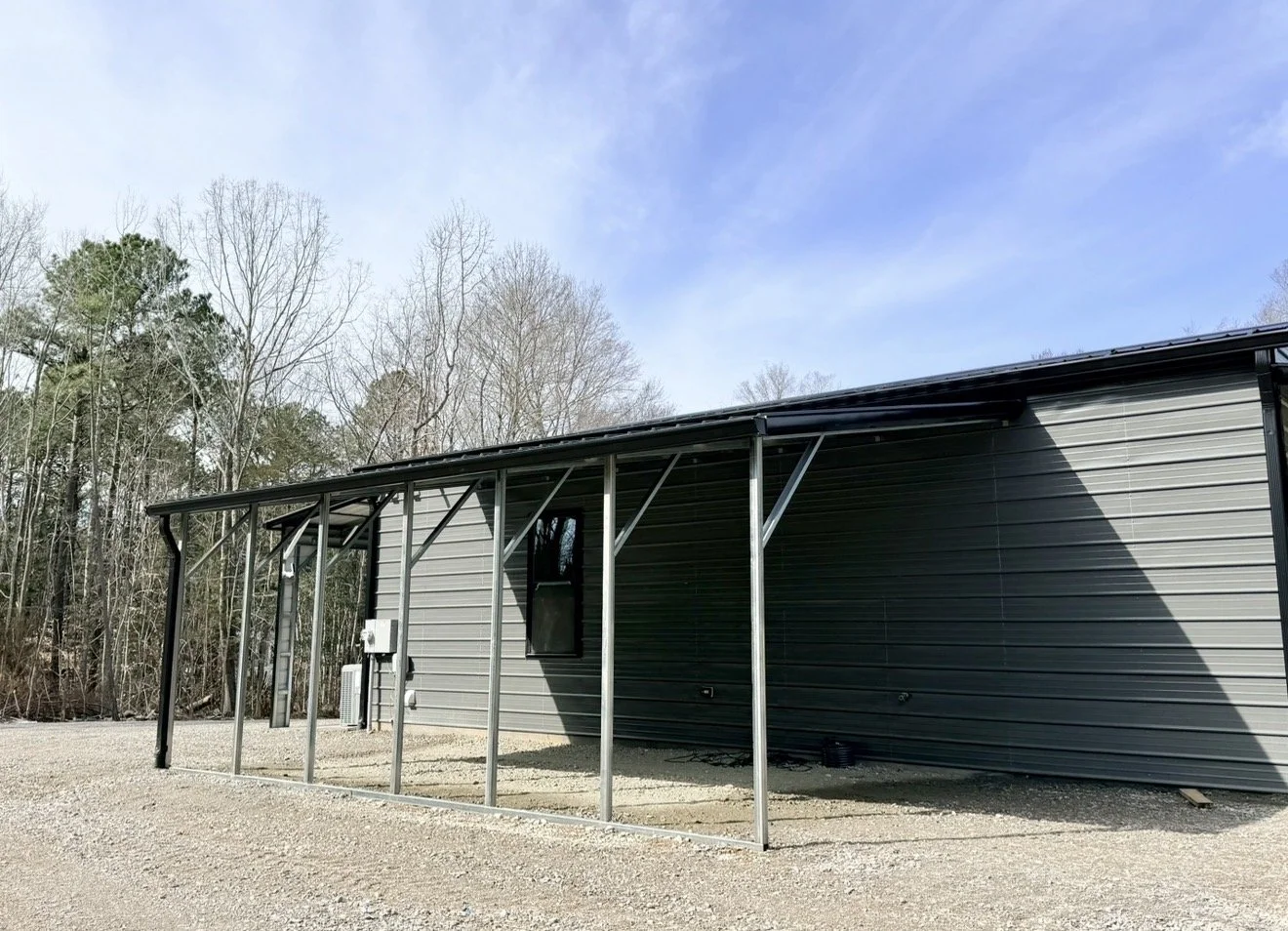 Newly constructed building with metal siding and a partial metal canopy, situated in a rural area with leafless trees and clear sky in the background.