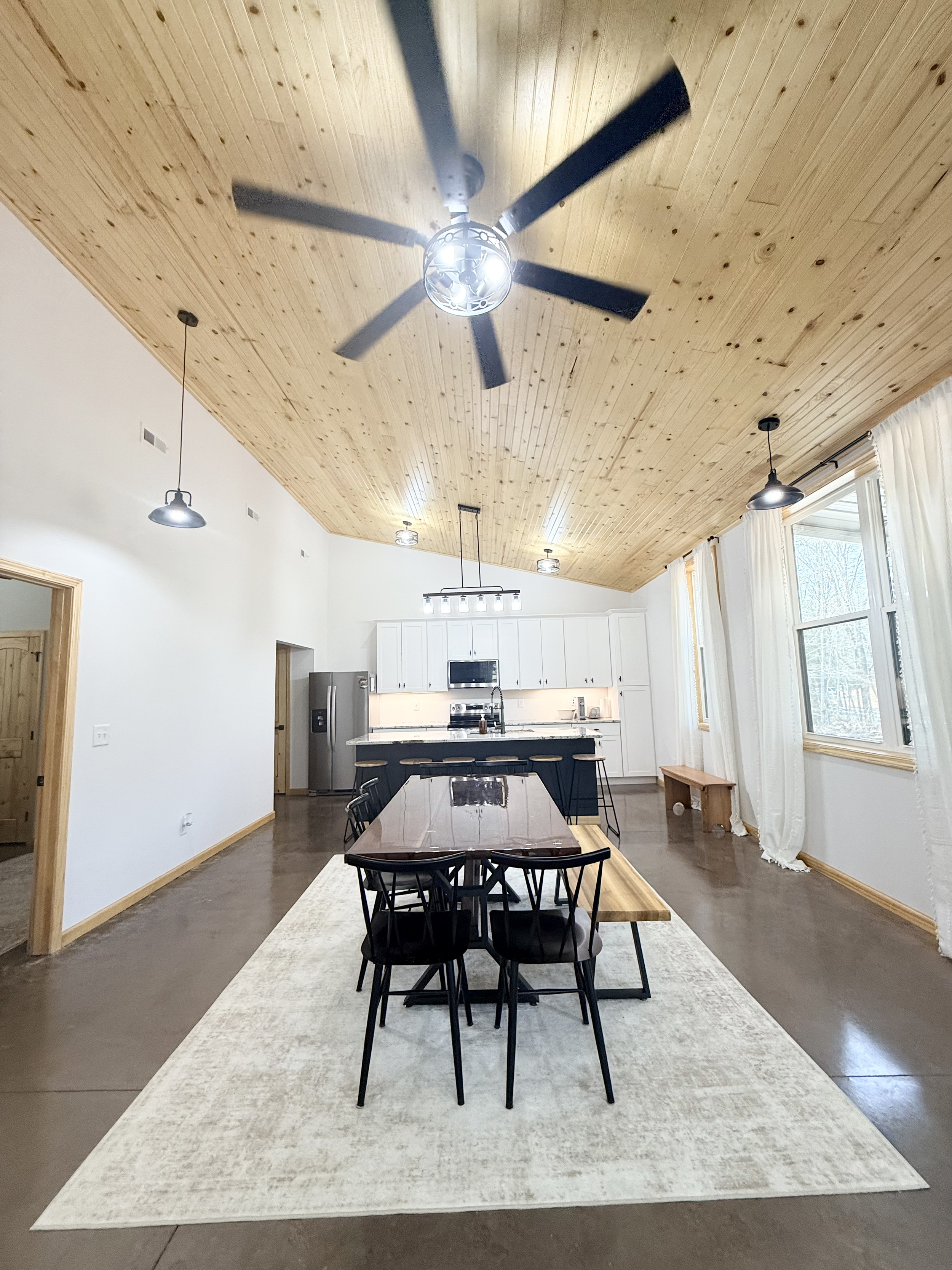 A spacious dining area with a dark wood table, black chairs, and a bench on a light-colored rug. The room has a wooden ceiling with a ceiling fan and multiple light fixtures, large windows with white curtains, and a modern kitchen with white cabinets