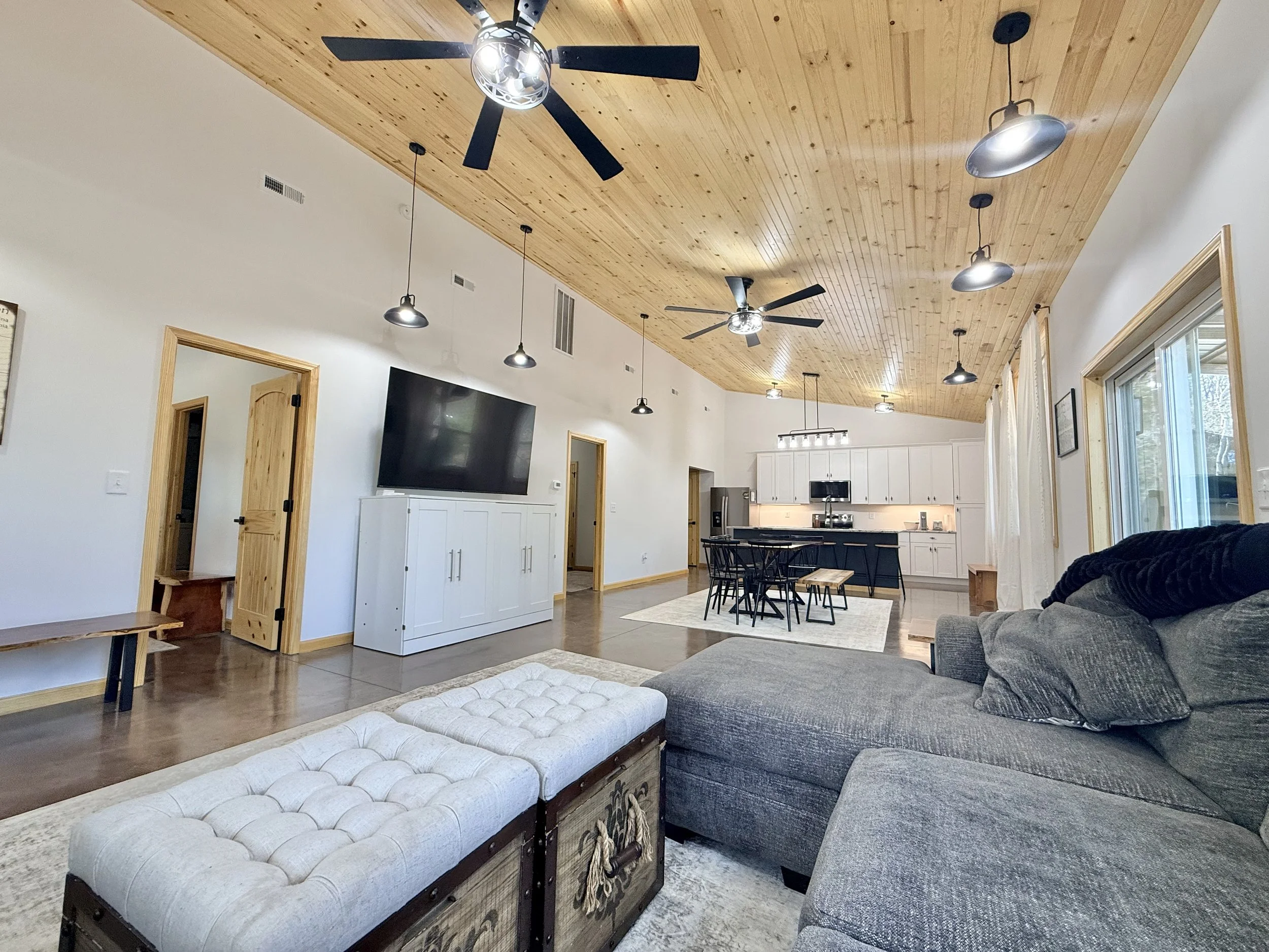 Living room with a wood-paneled ceiling, ceiling fans, pendant lights, a large gray sectional sofa, a tufted ottoman, a TV on a white cabinet, and an open kitchen with white cabinets and a black island.