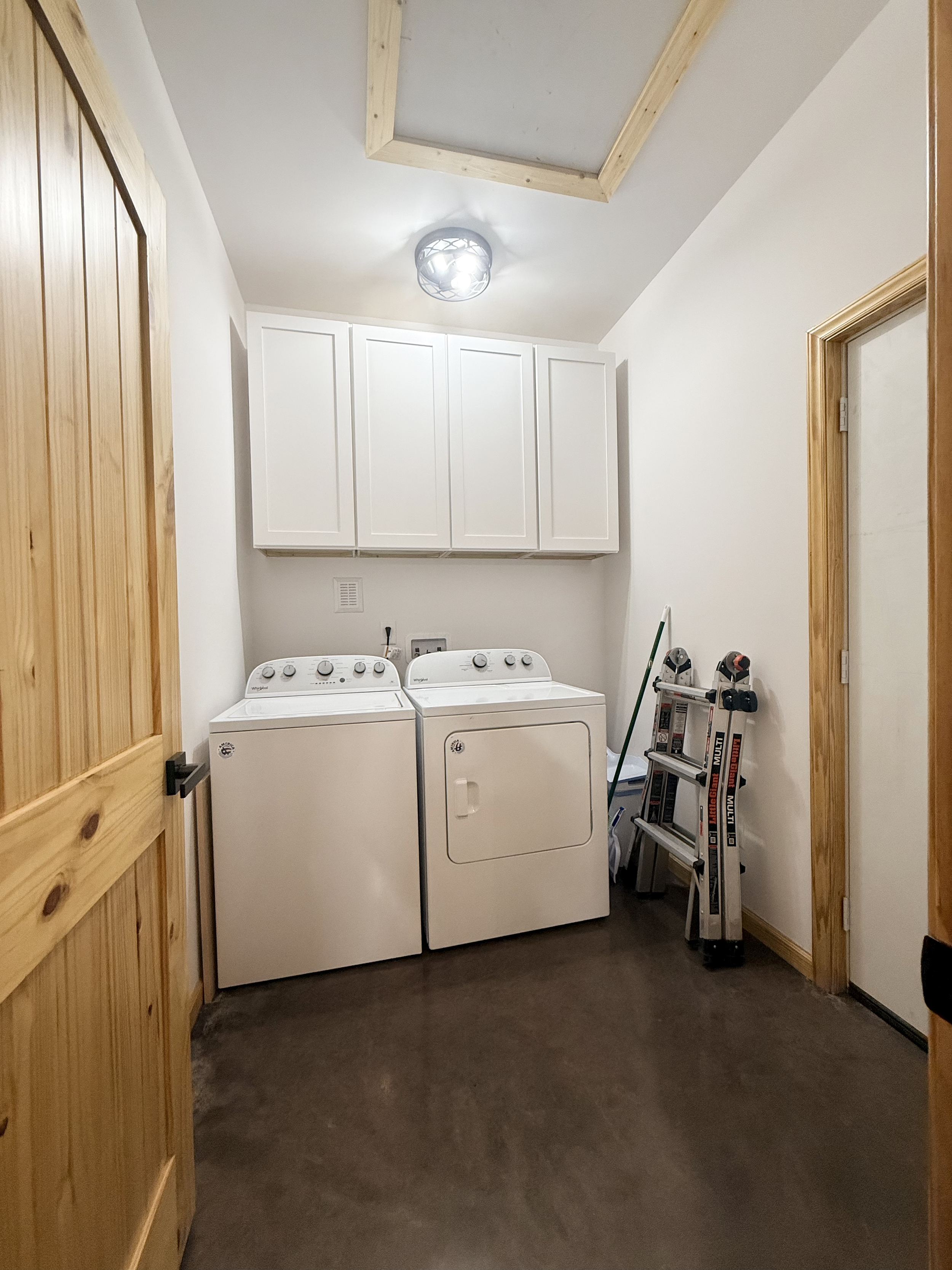 Laundry room with white washing machine and dryer, wooden door and cabinets, ladder, and cleaning supplies.