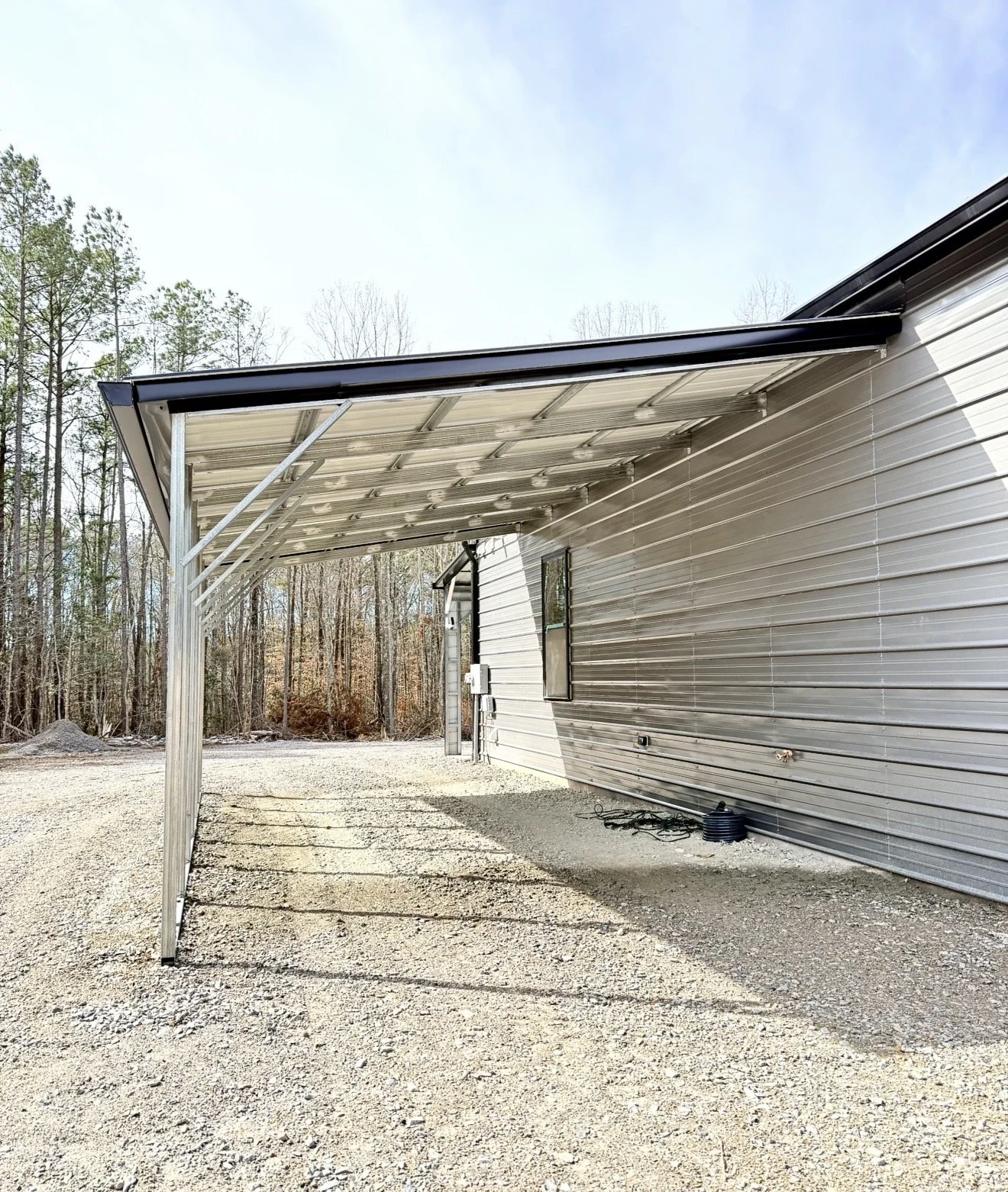Metal carport attached to a house with gravel driveway and trees in the background.