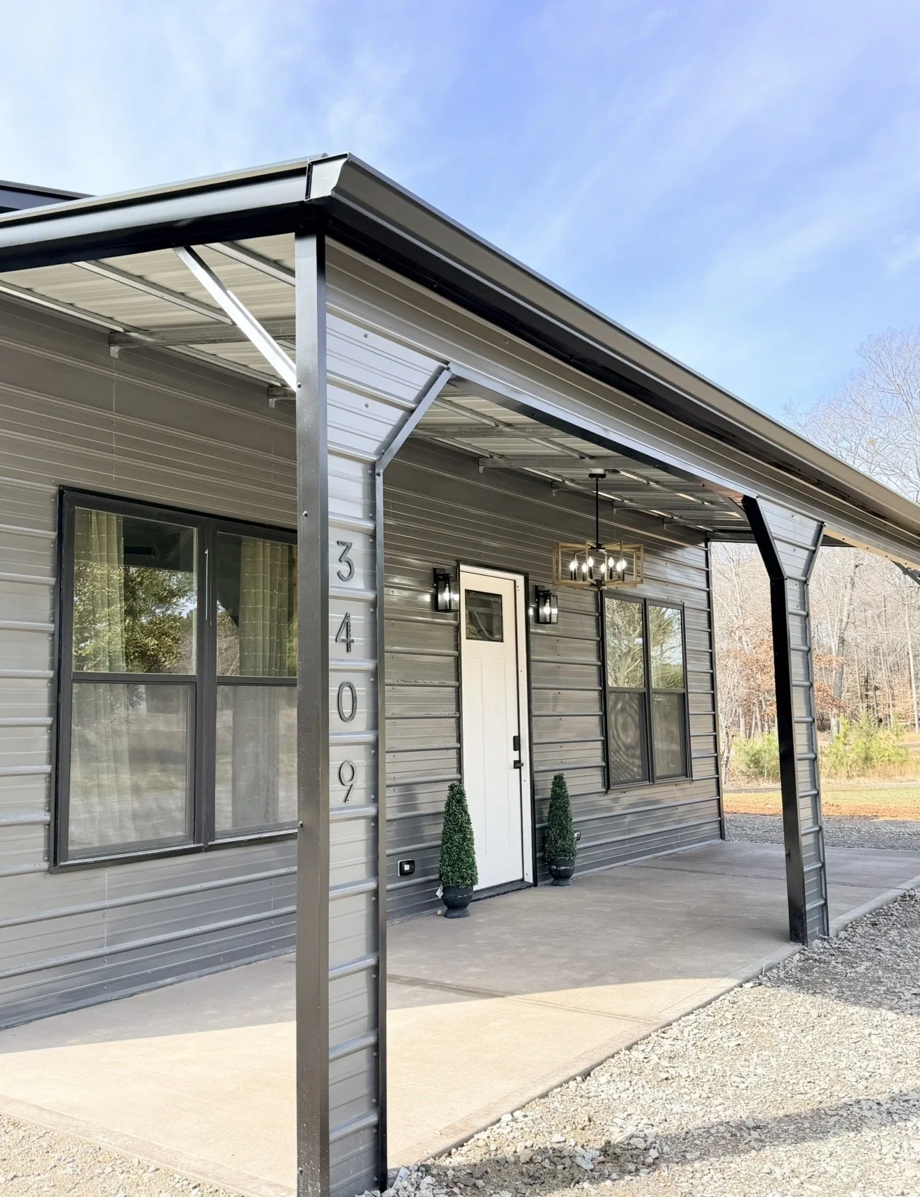 Front exterior of a modern metal house with a small porch, black-framed windows, and a white front door, decorated with two small potted evergreen plants, under a metal awning, with a blue sky and trees in the background.