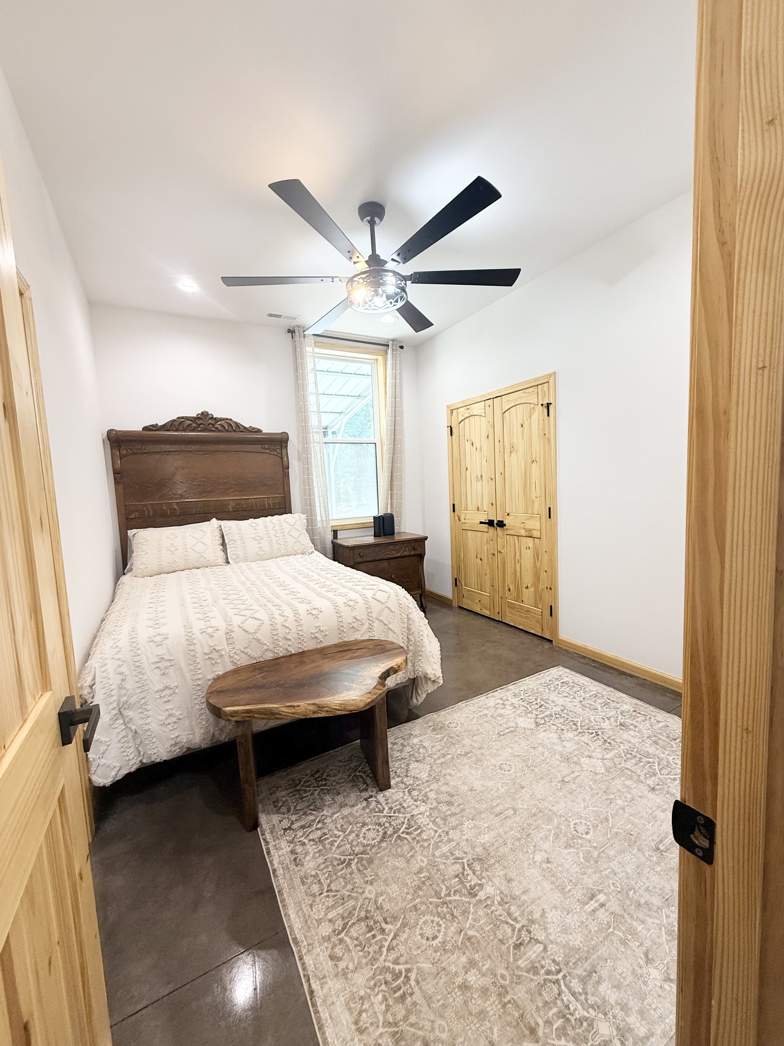 A bedroom with a wooden bed frame, white bedding, a black throw blanket, and a wooden nightstand. There is a window with light curtains, a patterned area rug, a wooden bench at the foot of the bed, and a ceiling fan with five blades. The room has white walls and wooden trim.