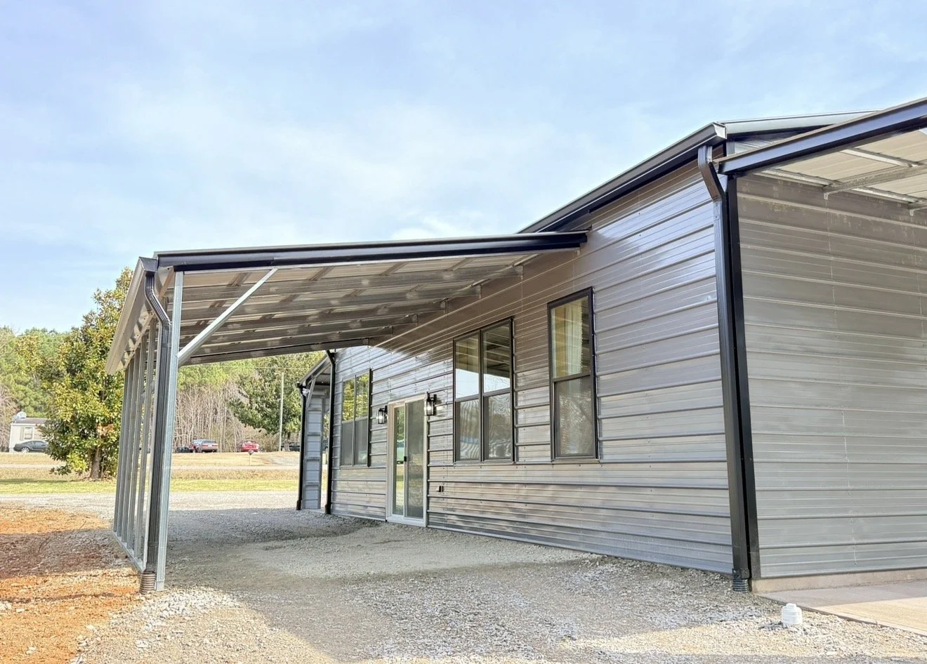 Exterior of a metal building with a small covered area, gravel ground, and trees in the background.