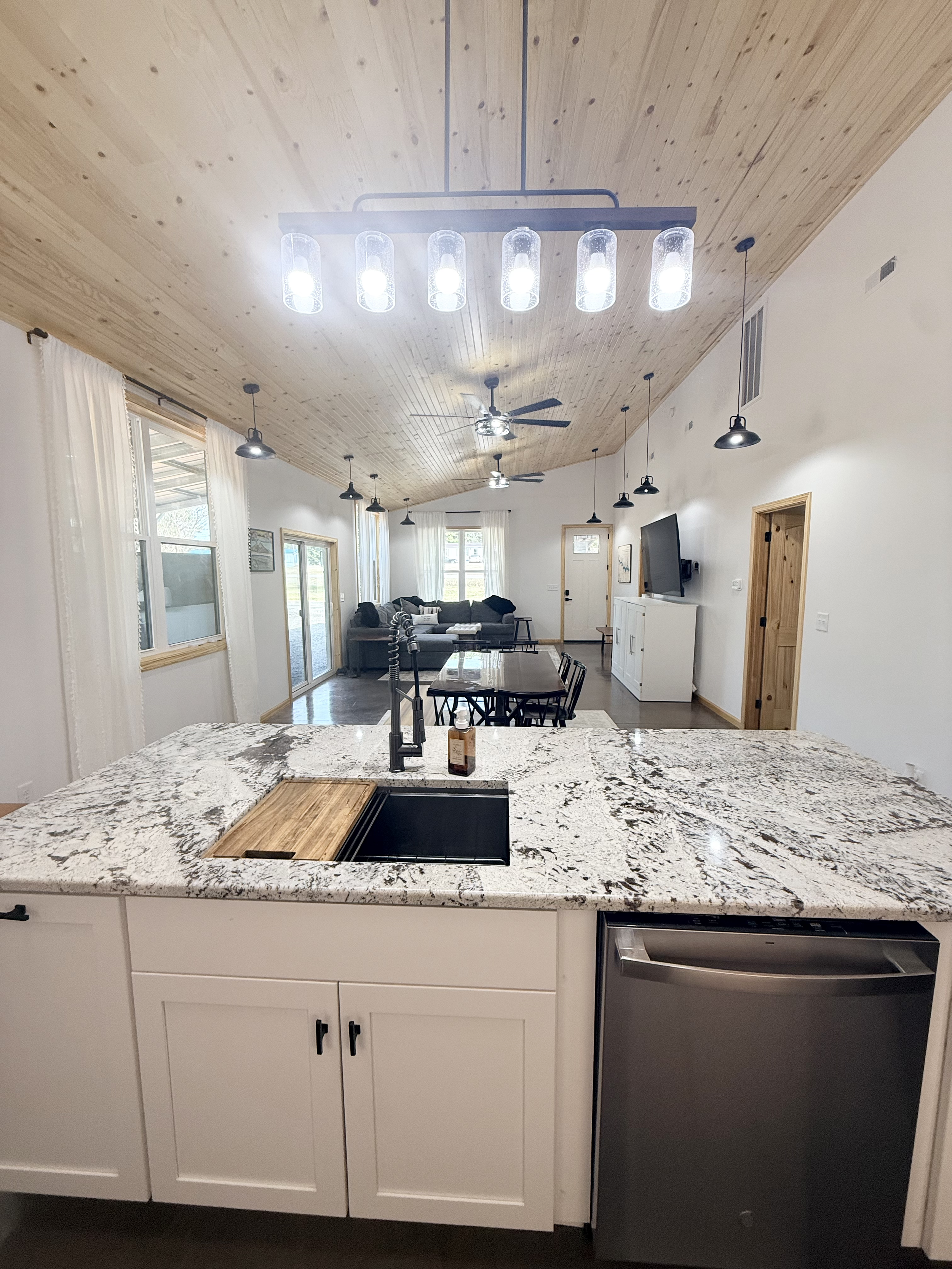 Modern open-concept kitchen with granite countertops, white cabinets, and a black sink, leading into a living and dining area with wooden ceiling, ceiling fans, and large windows.