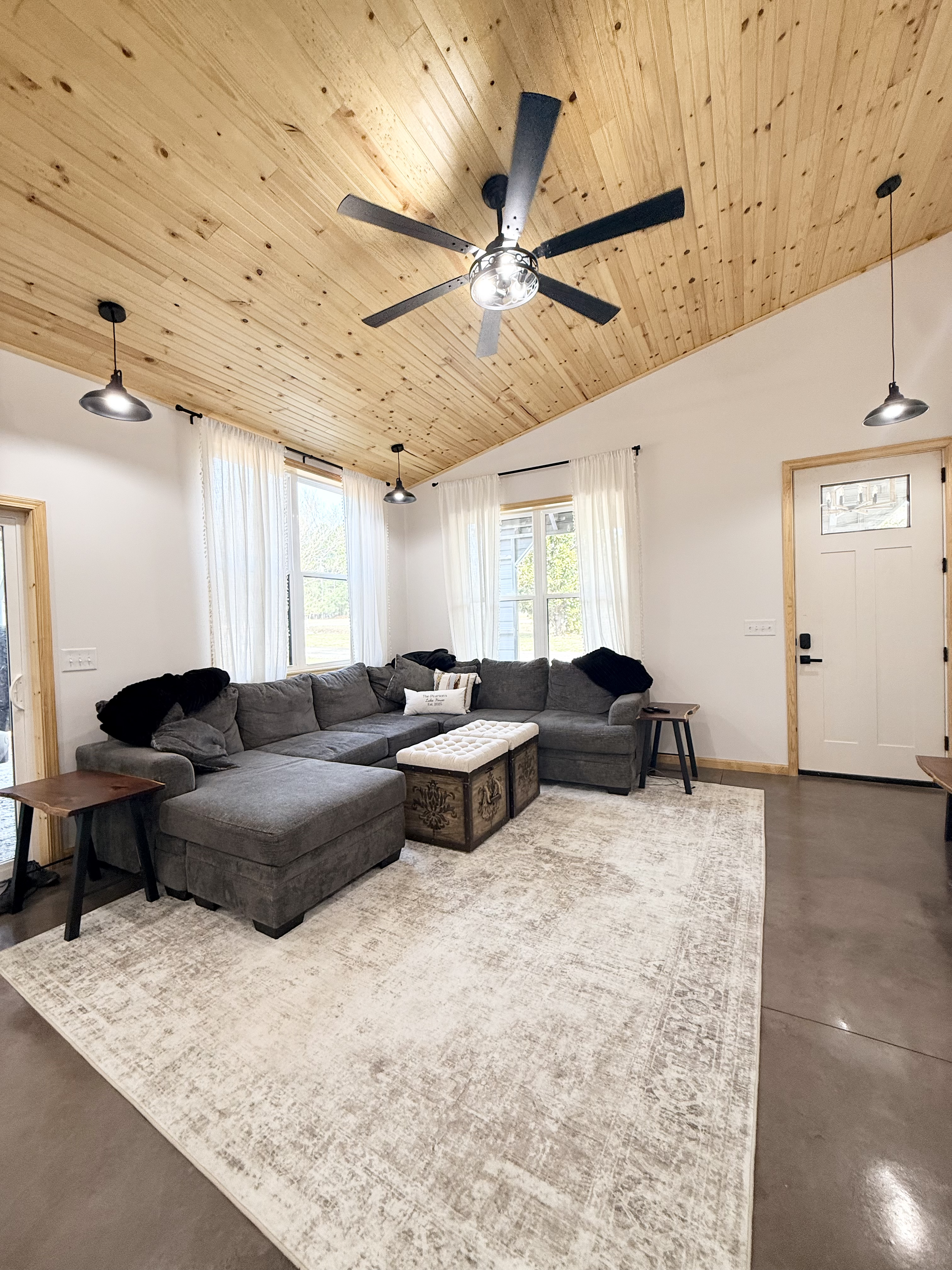 Living room with gray sectional sofa, wooden ceiling, white walls, windows with sheer curtains, ceiling fan, and a light-colored area rug.