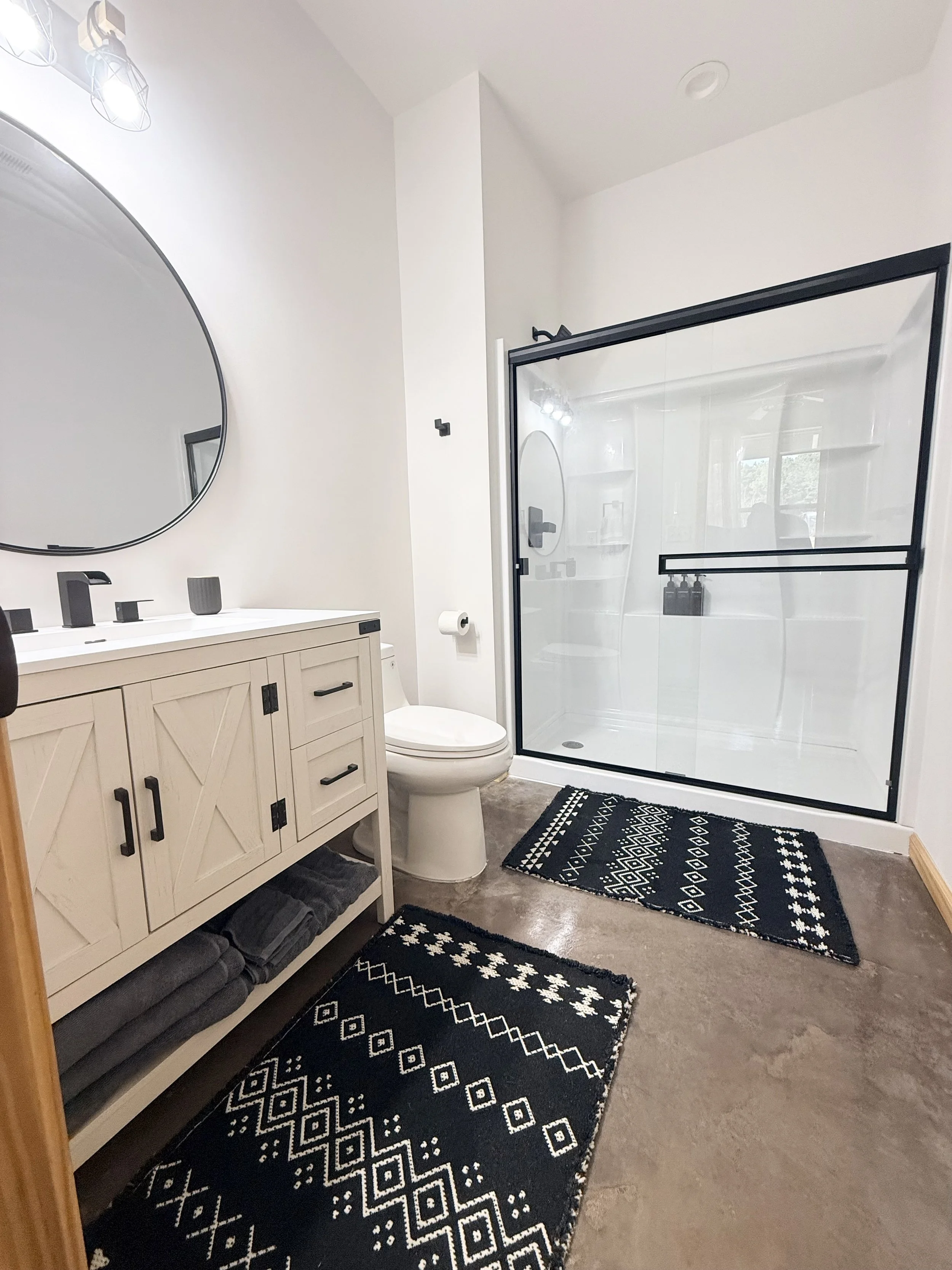 Modern bathroom with a white vanity, round mirror, black fixtures, toilet, black and white patterned rugs, and a glass shower enclosure with a black frame.