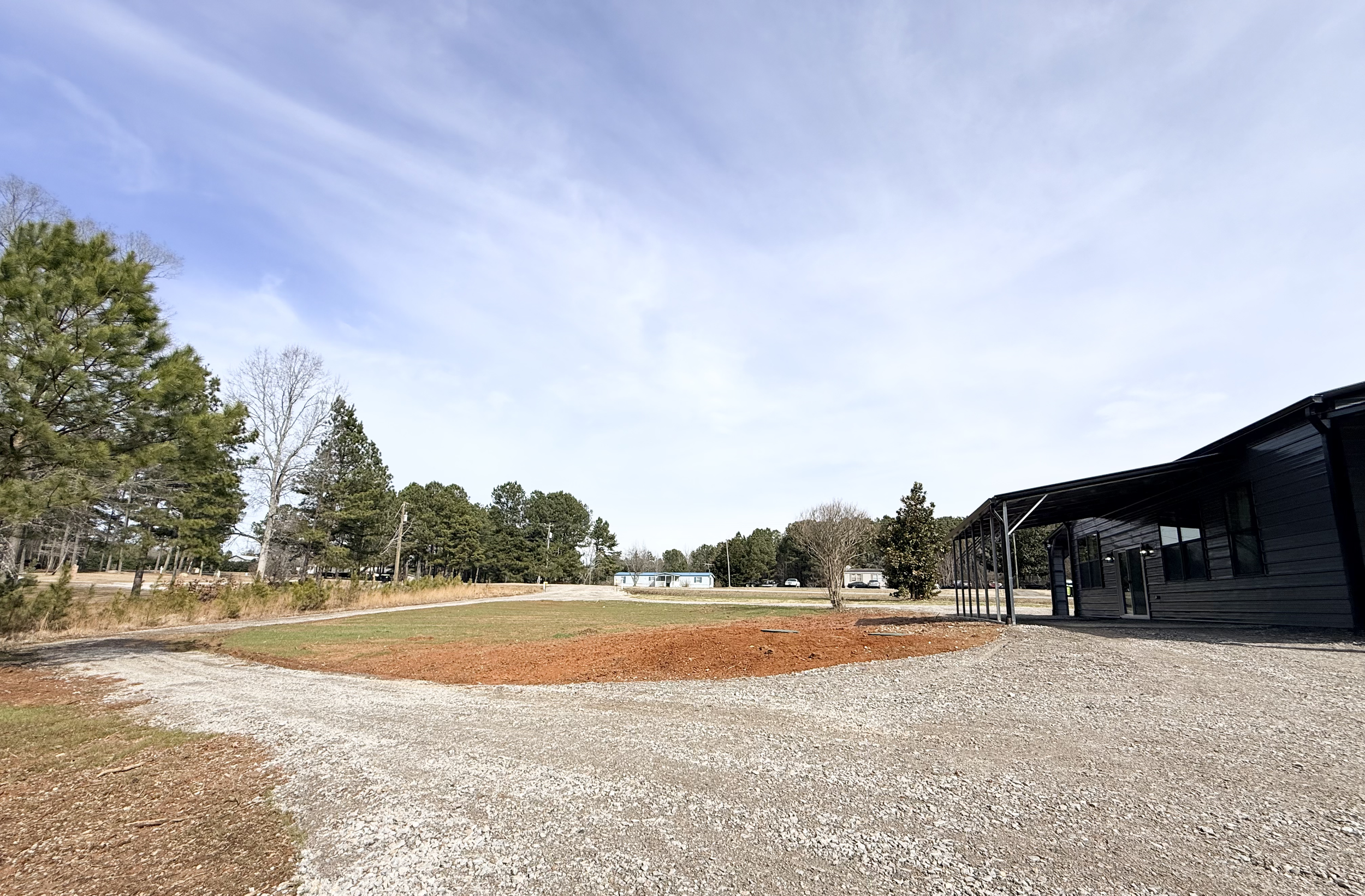 Exterior of a black modern building with a gravel path and a small grass area, surrounded by trees under a partly cloudy sky.
