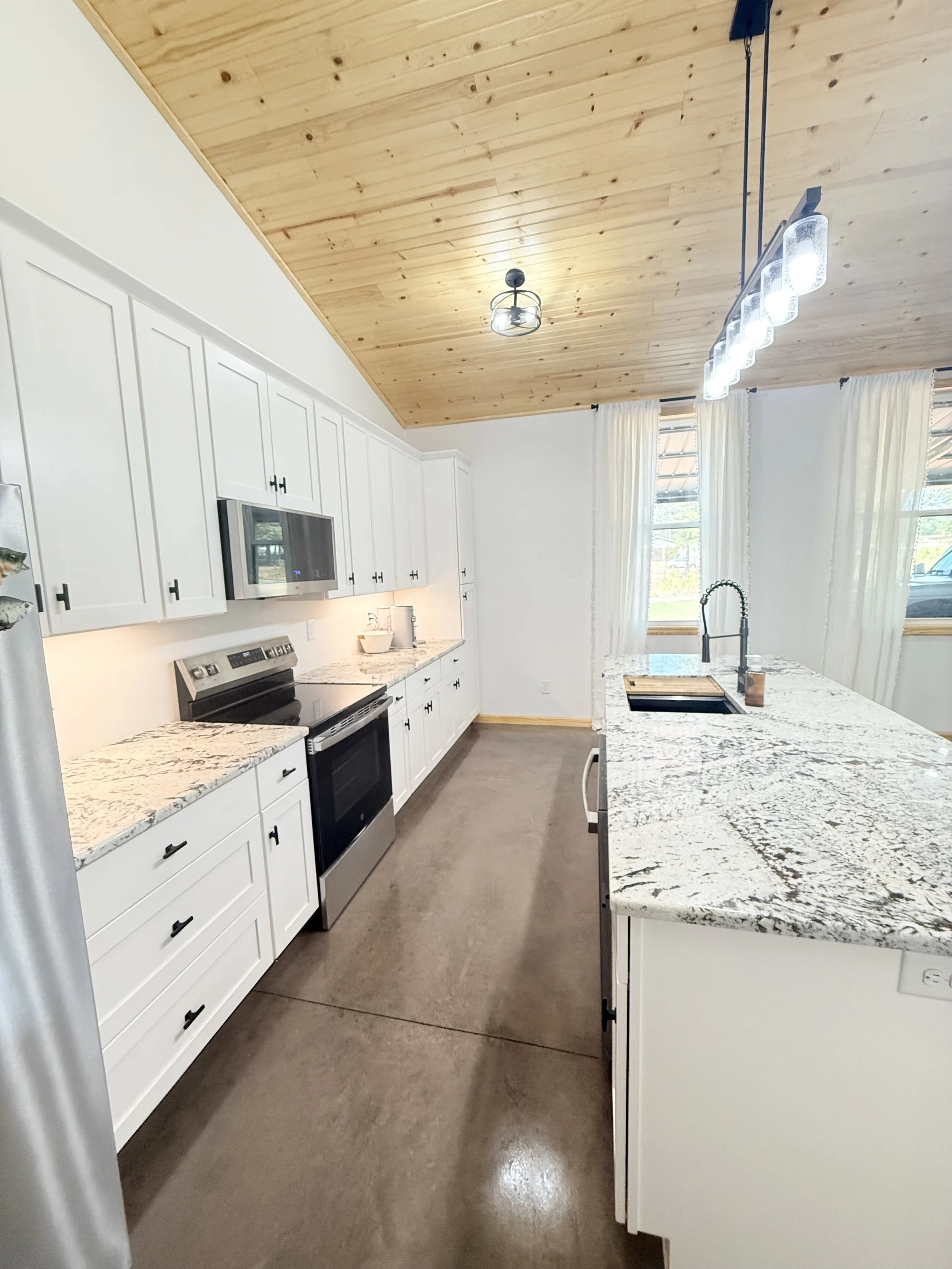 Modern kitchen with a large island featuring a patterned granite countertop, surrounded by six stools with wooden seats and black metal legs. White cabinets, stainless steel appliances, and a small window with curtains are visible in the background.
