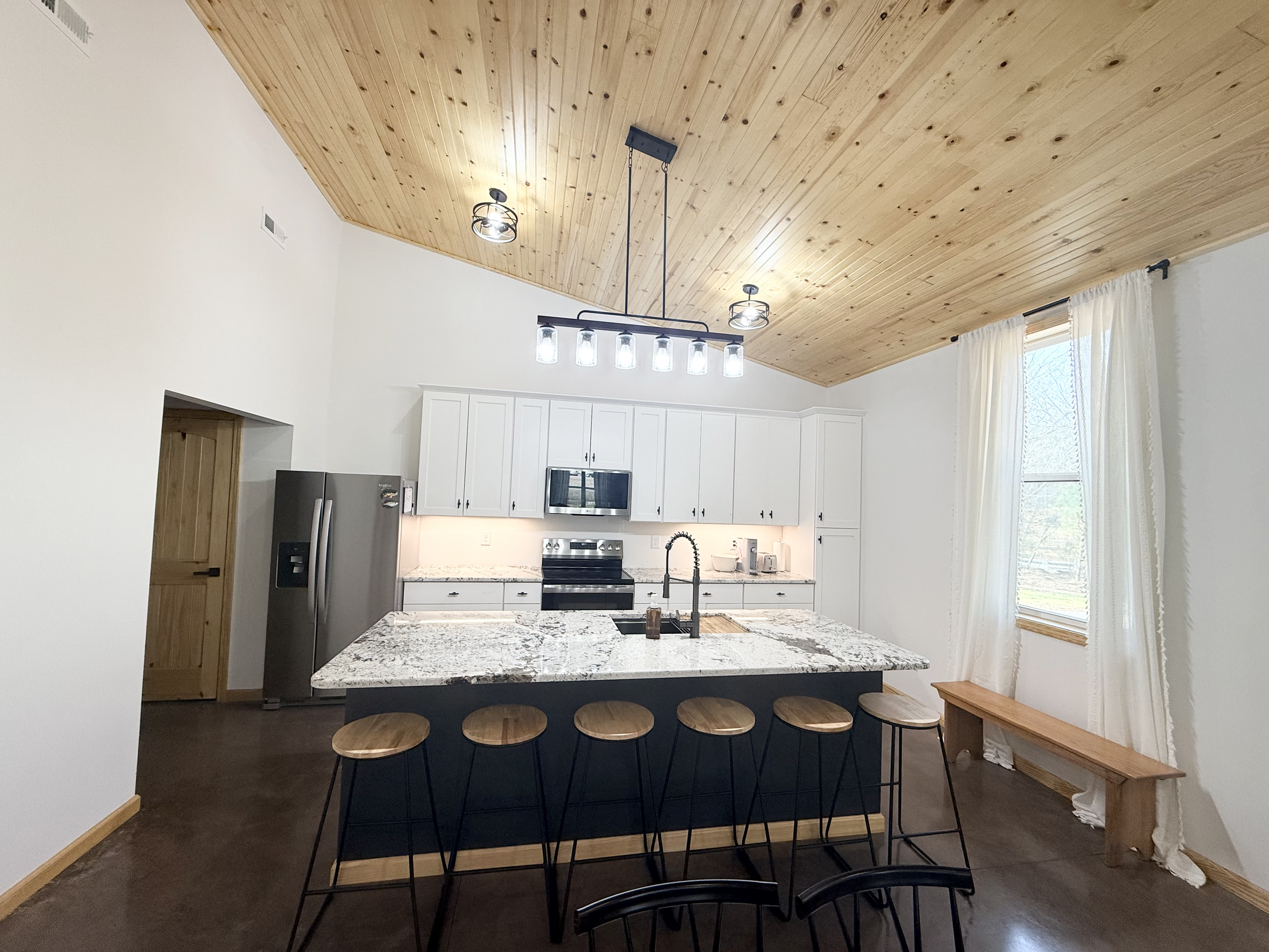 Modern kitchen with white cabinets, granite countertops, stainless steel appliances, a large island with a granite top and wooden bar stools, wooden ceiling, and window with white curtains.