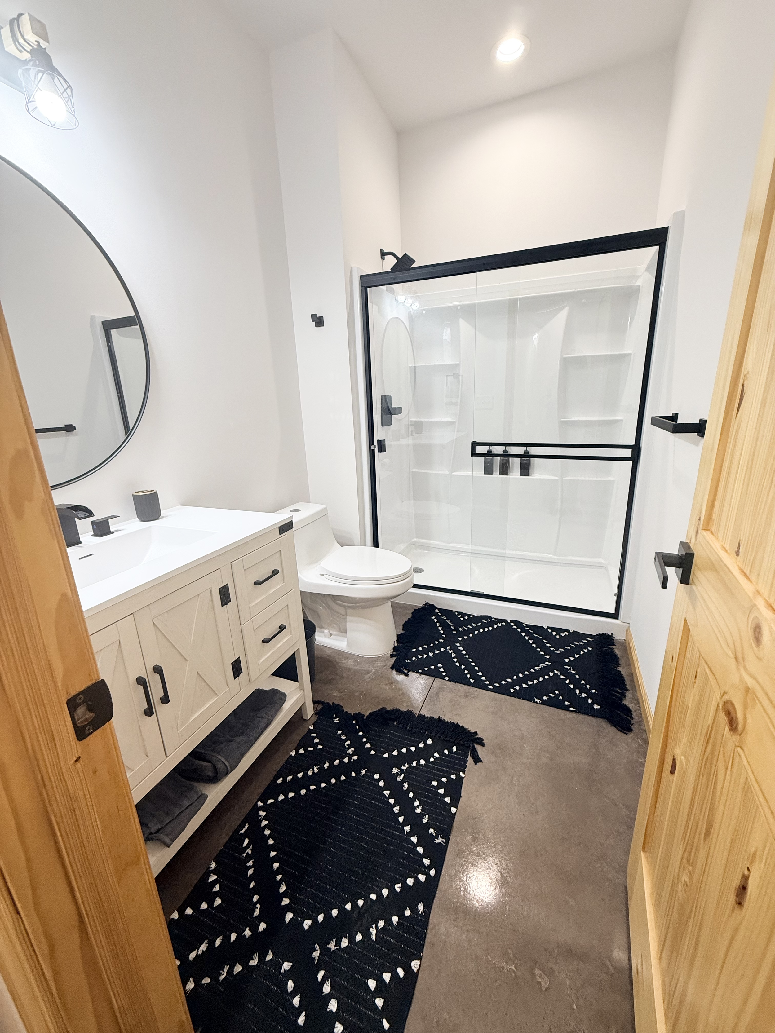 A modern bathroom with a white vanity and round mirror, a white toilet, a glass shower with a black frame, black towel bars, black soap dispensers, and two black patterned rugs.