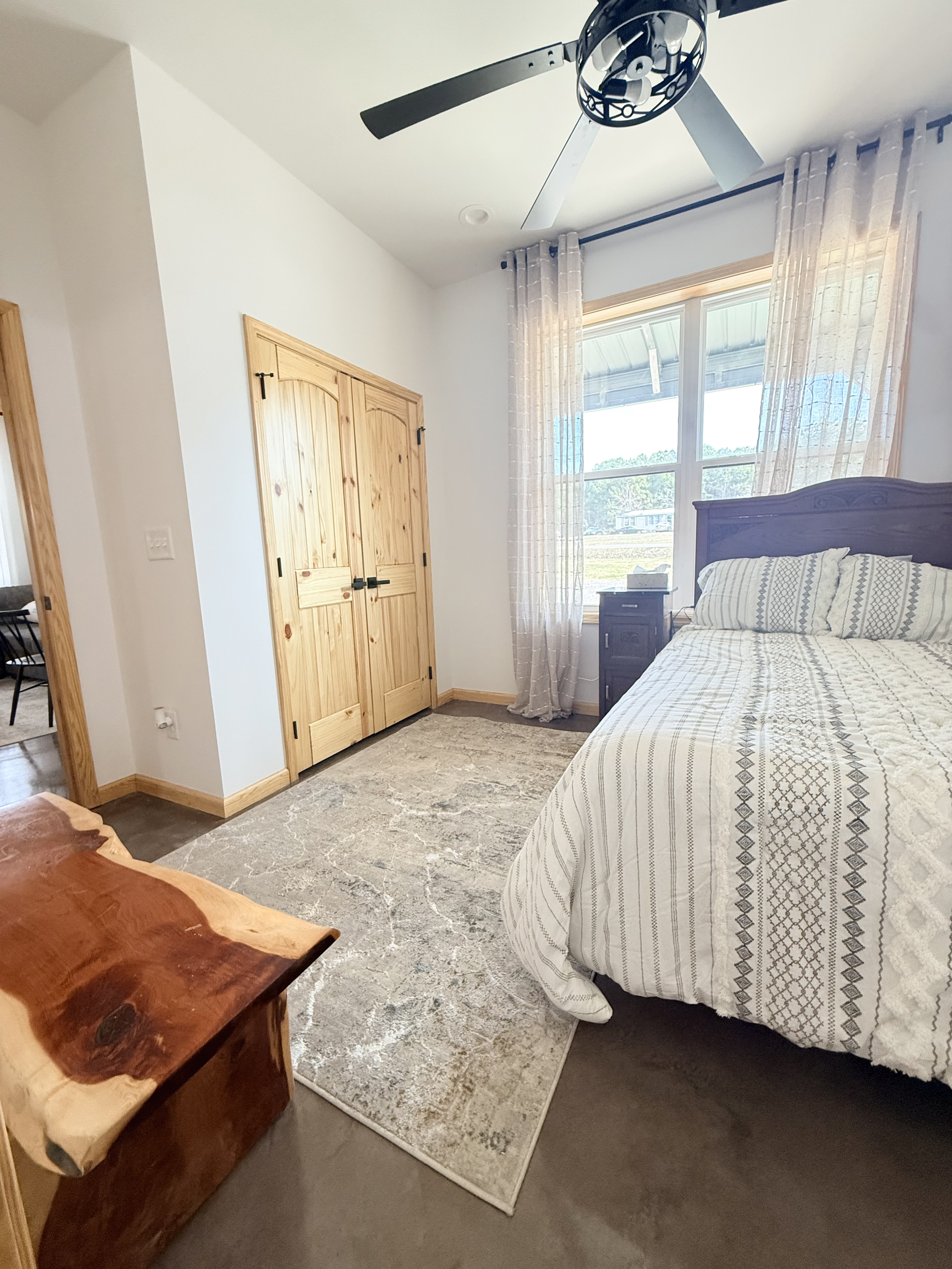 A bedroom with a large window, wooden closet doors, a bed with decorative bedding, a nightstand with a clock, a ceiling fan, and a carpeted floor.