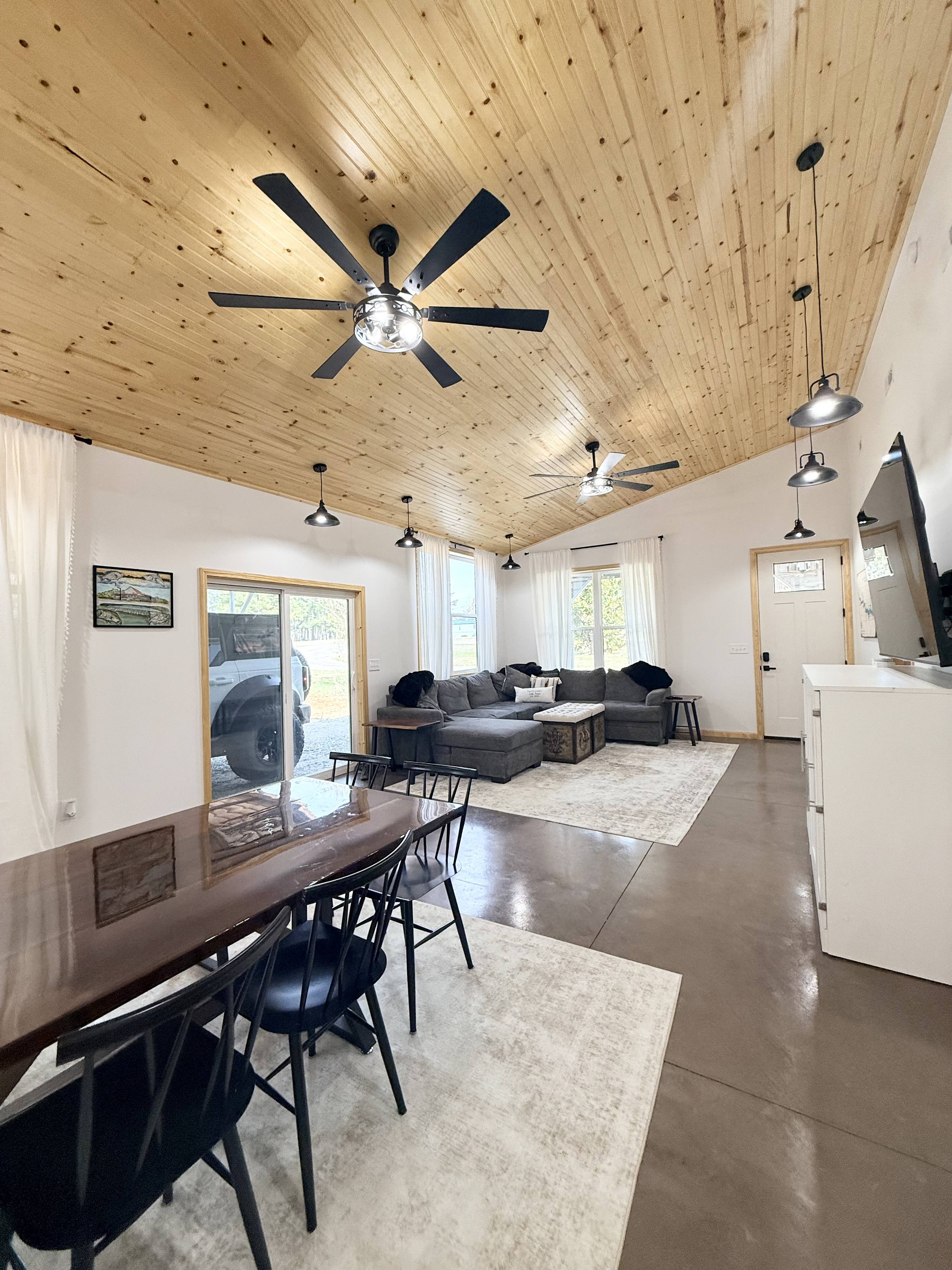 Living room with a wooden ceiling, ceiling fans, pendant lights, white walls, large windows with white curtains, a gray sectional sofa, a wooden chest, a dark wooden dining table with black chairs, a white cabinet, and wall art.