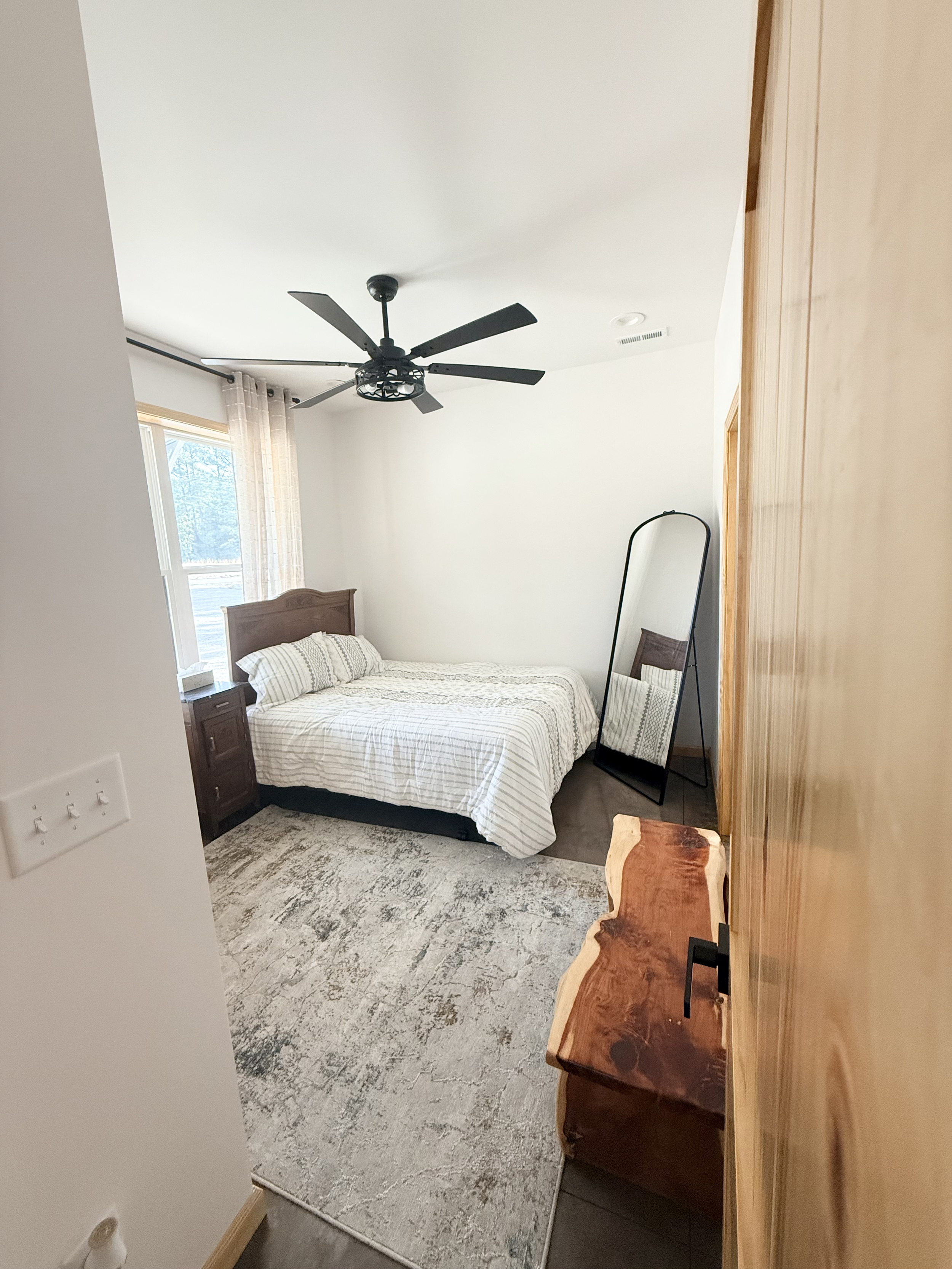 Bedroom with a bed, window, standing mirror, wooden nightstand, wooden dresser, ceiling fan, and a rug.
