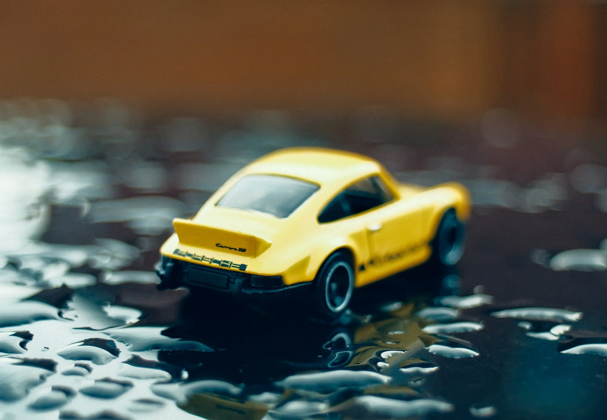 Yellow toy Porsche car on a wet reflective surface with blurred background.