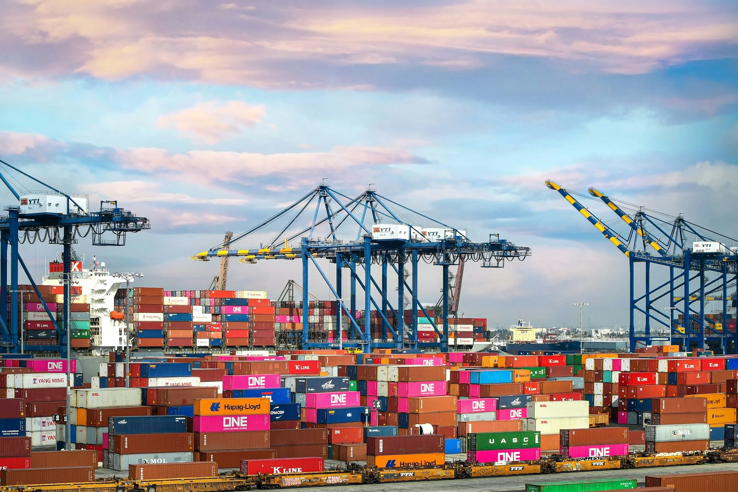 View of a shipping port showing multiple cargo containers, large blue cranes, and a cargo ship in the background under a partly cloudy sky.