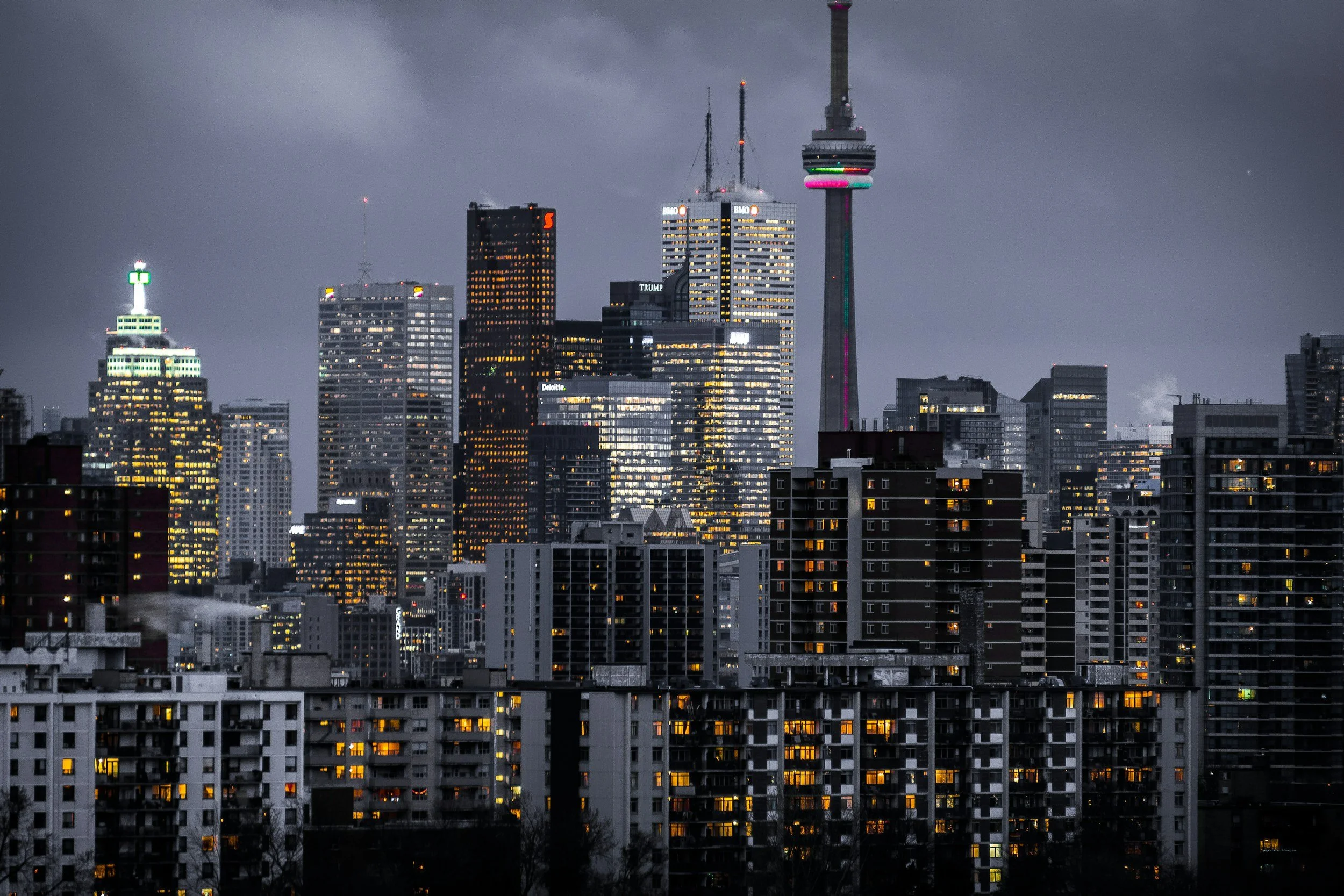 Nighttime city skyline with skyscrapers, some with lit windows. Tower with colorful lights on the right and a green-lit building on the left, overcast sky.