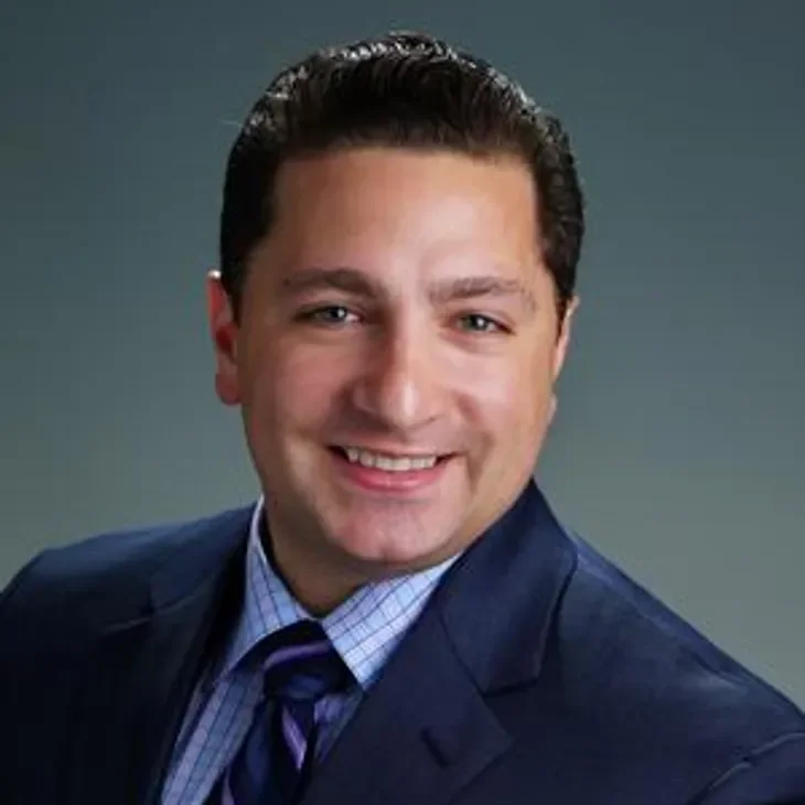 A professional headshot of a smiling man with dark hair, wearing a dark suit, light blue dress shirt, and a striped tie, against a gray background.