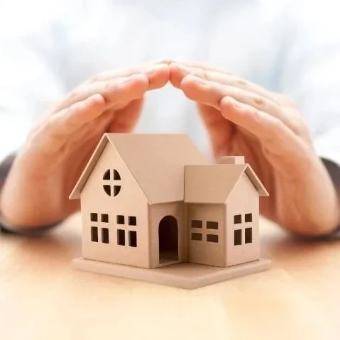 Hands protecting a small cardboard house placed on a wooden surface.