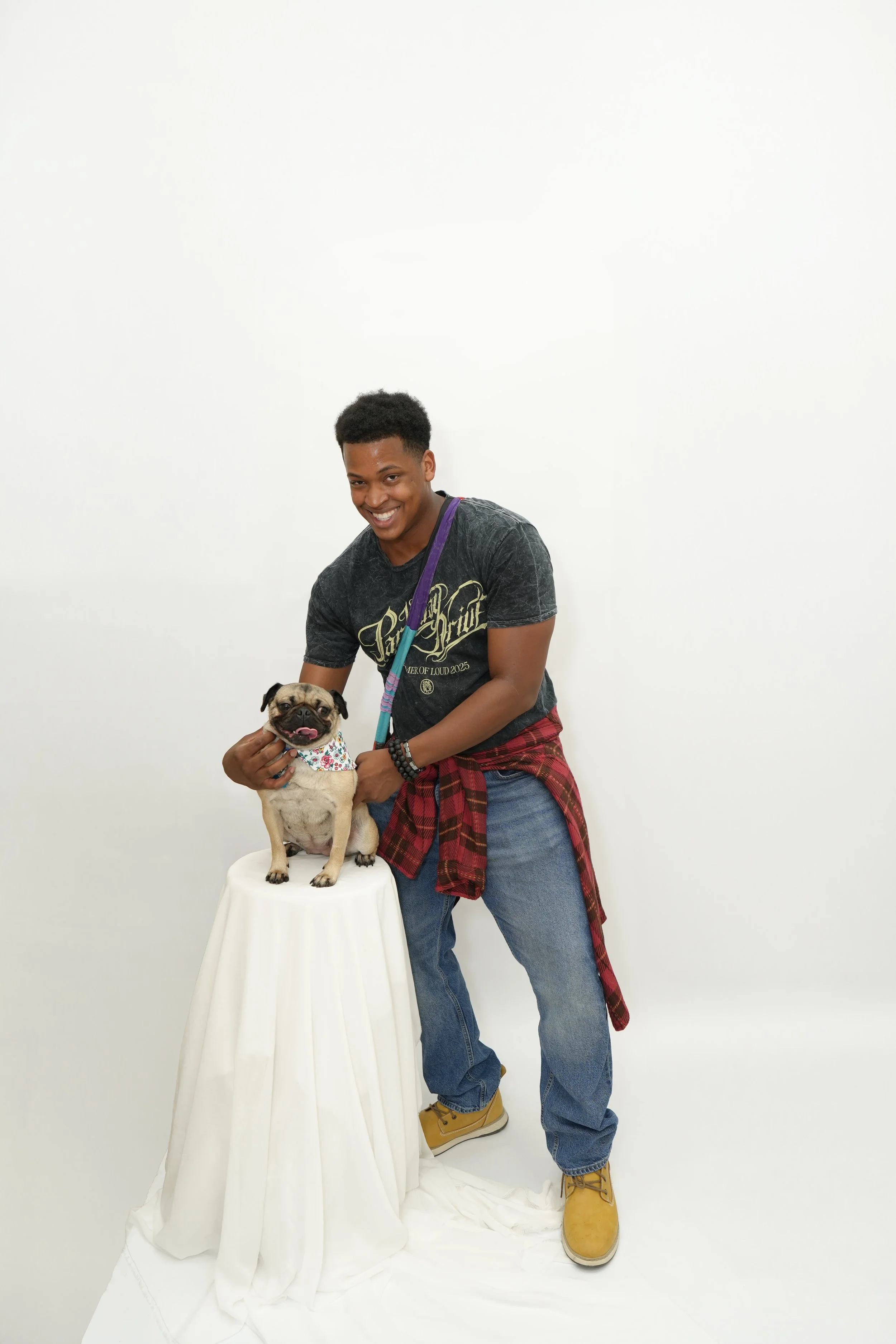 A young man with a smile bending to hold a small pug dog on a white table, standing against a plain white background. The man is wearing a black graphic t-shirt, blue jeans, yellow boots, and has a plaid shirt tied around his waist. The dog is wearin