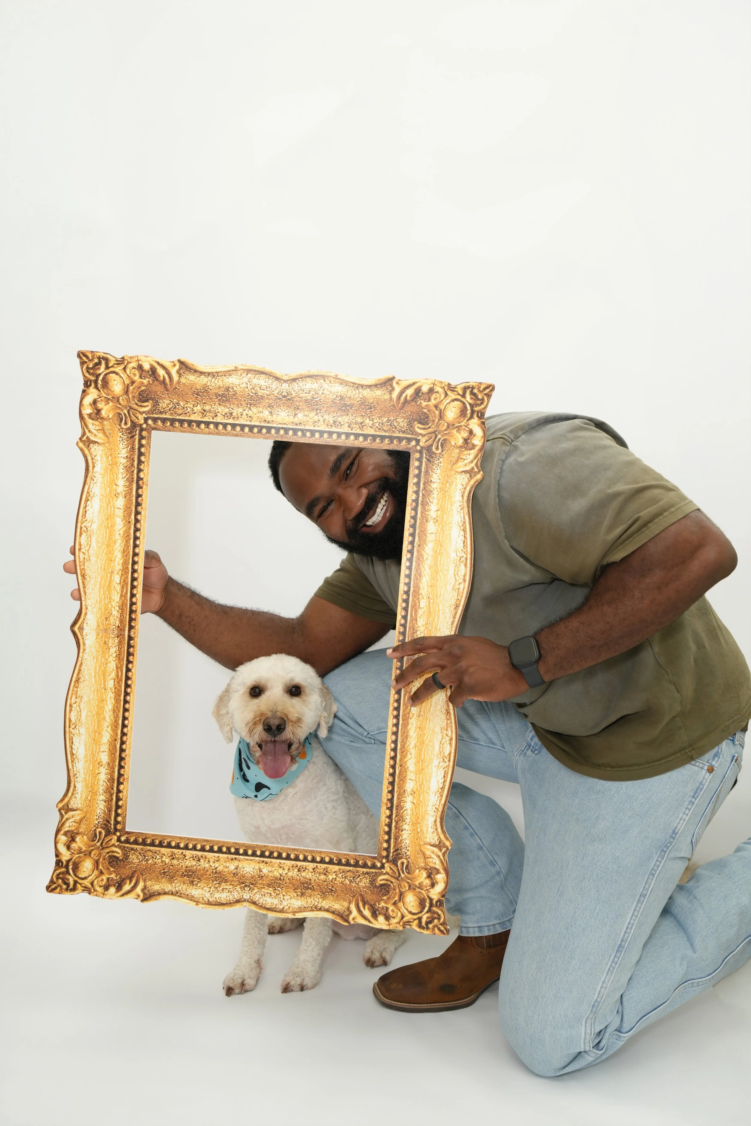 A man with a beard, smiling, holding a large ornate gold picture frame while kneeling next to a white dog with curly fur. The dog is wearing a blue bandana with black spots and has its tongue out. The man is dressed in a green T-shirt, light blue jea