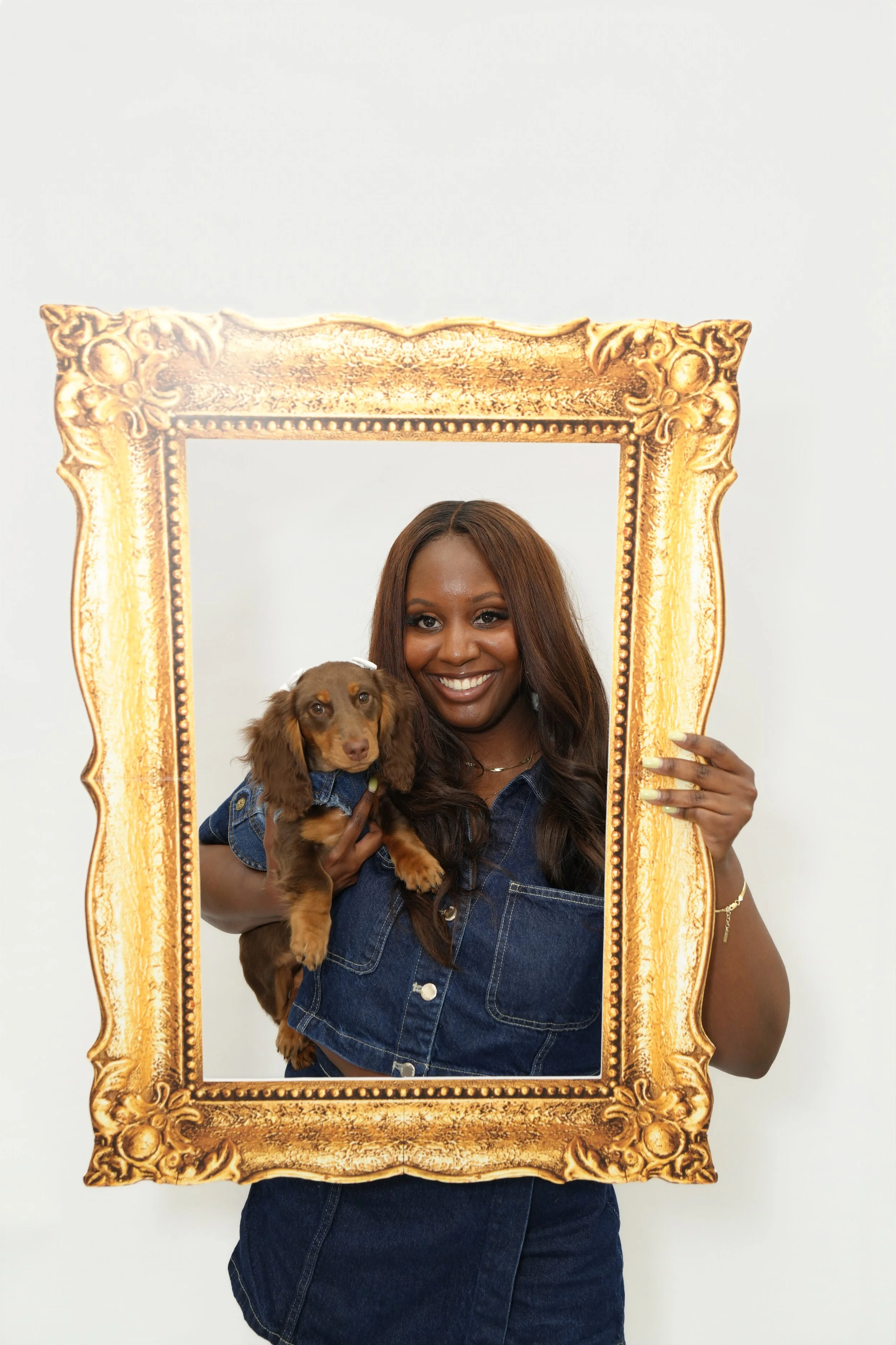 A woman with long brown hair is smiling and holding a brown dog with a denim jacket inside a large ornate gold picture frame, against a plain white background.