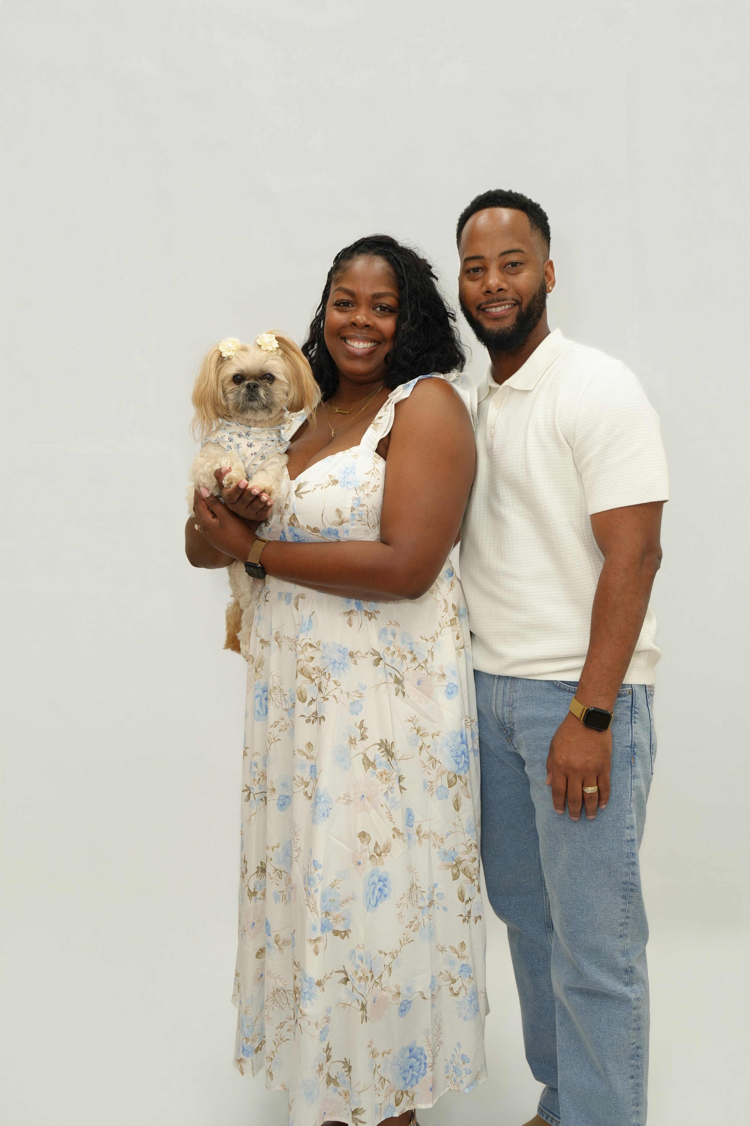 Group photo of a smiling Black woman holding a small dog with flower accessories on its head, standing next to a smiling Black man, against a plain white background.