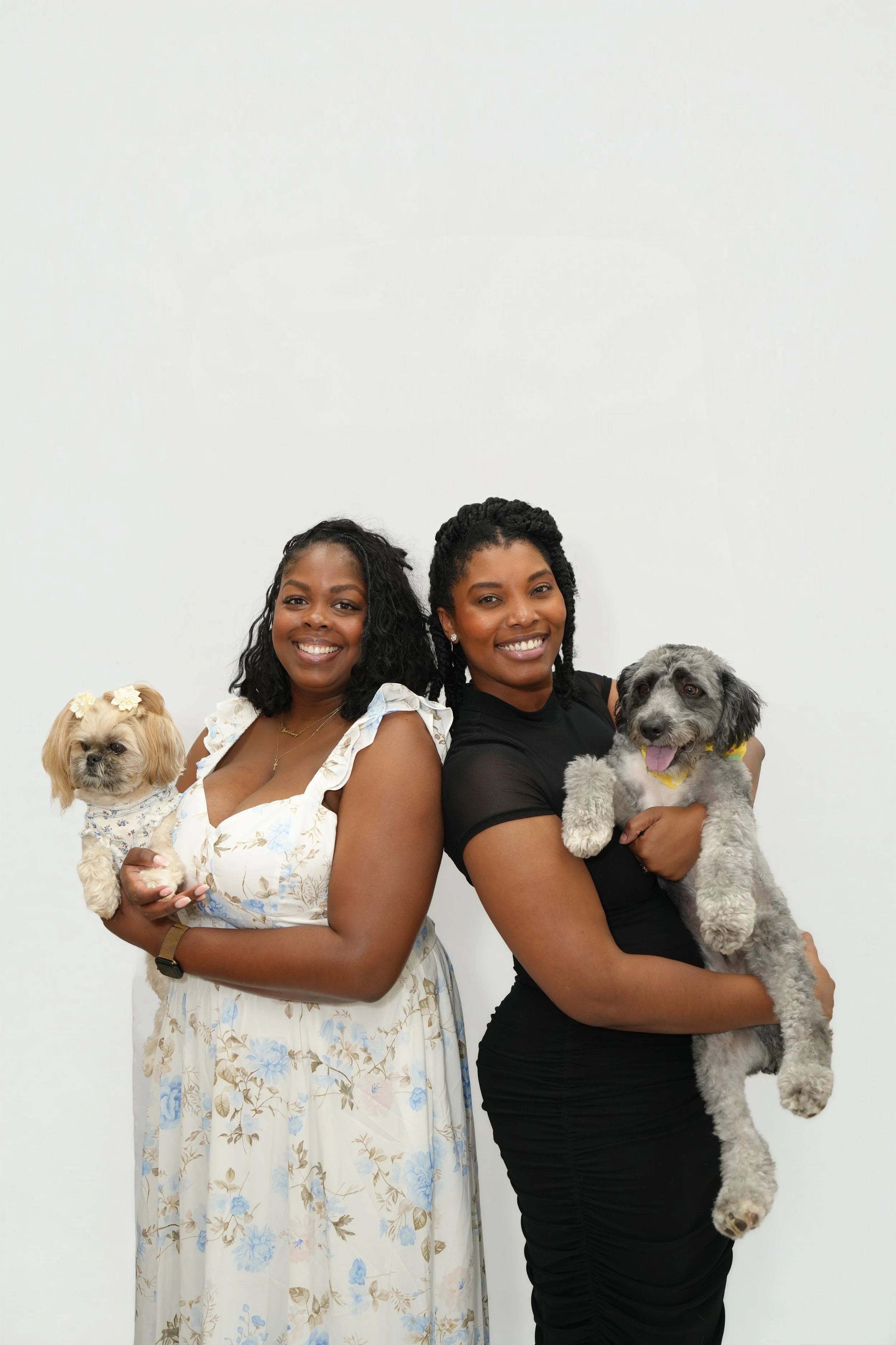 Two women standing against a plain white wall, each holding a dog, smiling at the camera.