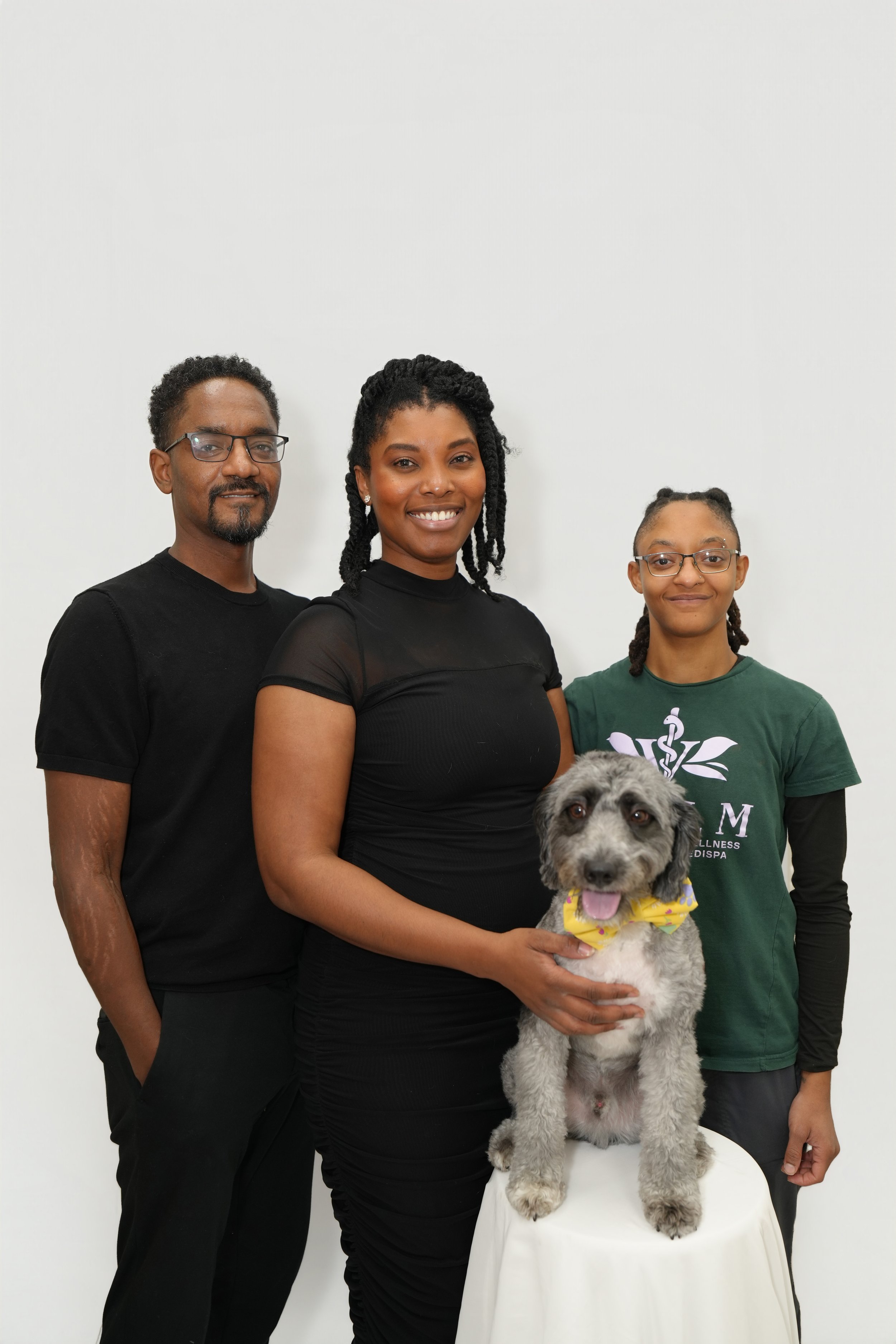 A group of four people, three women and one man, standing behind a small table with a dog on it. The woman in the center is holding the dog, a large mixed breed with curly gray fur and a colorful bandana around its neck. The group is smiling against 