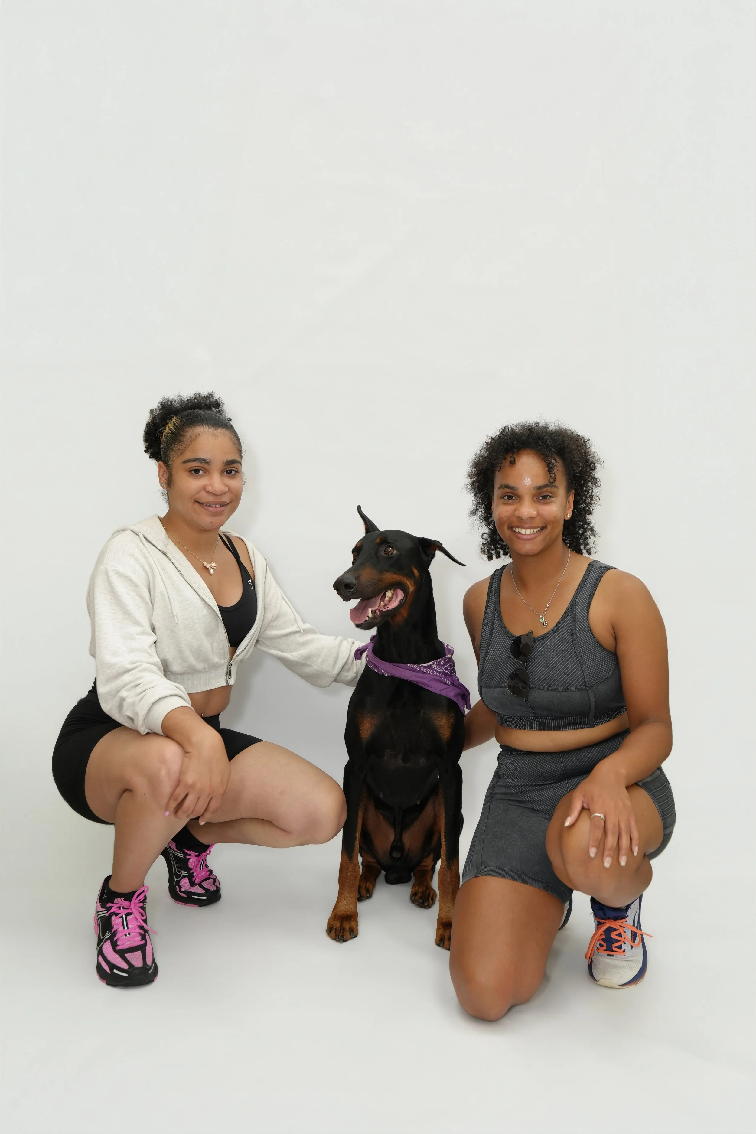 Two women and a Rottweiler dog posing together against a plain white background. The woman on the left is wearing a light-colored hoodie and black shorts with pink sneakers. The woman on the right is wearing a gray tank top and matching shorts, with 