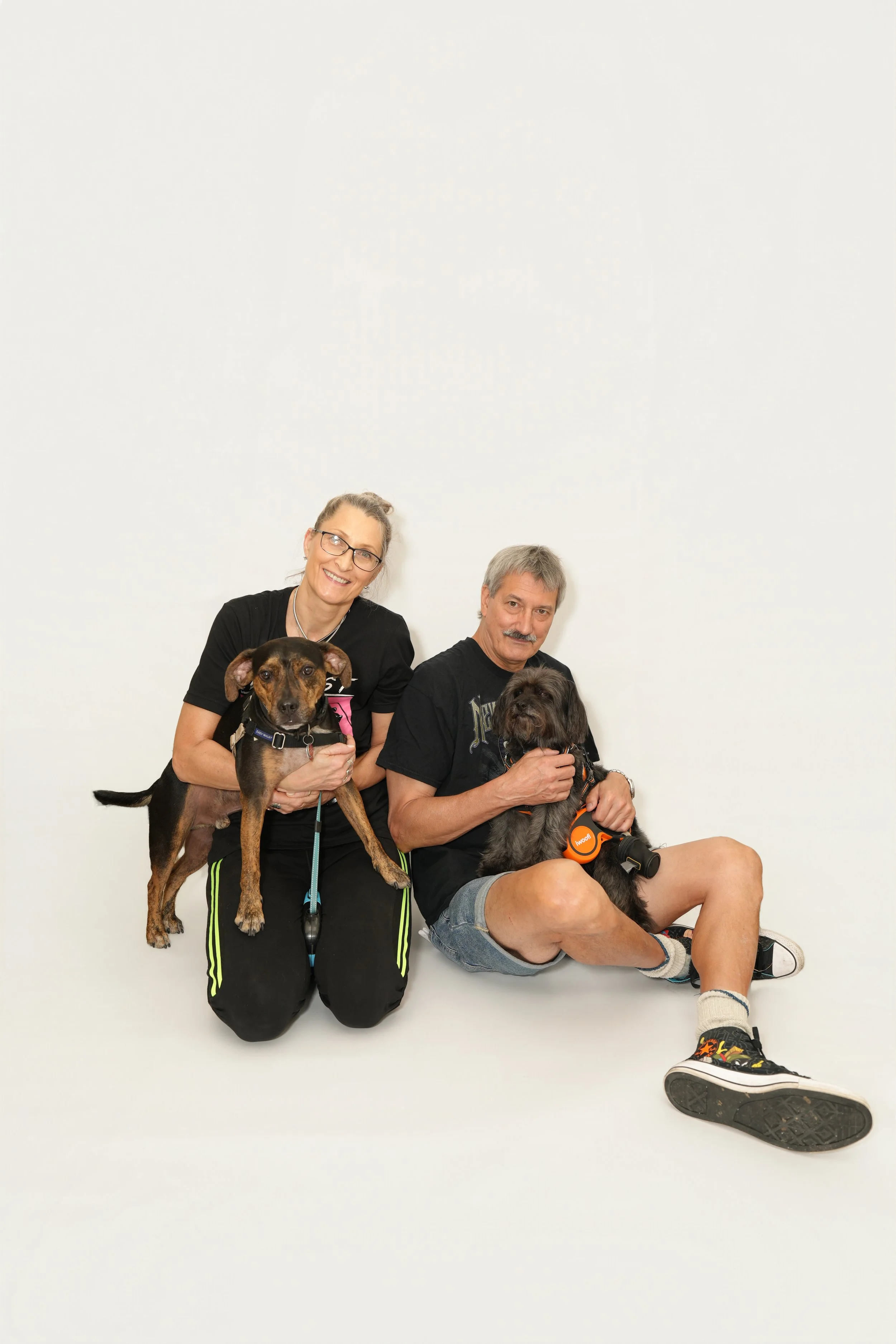 A smiling woman with glasses and a man with a mustache holding a small dog, both sitting on the floor against a plain white wall, with a large dog standing in front of the woman.