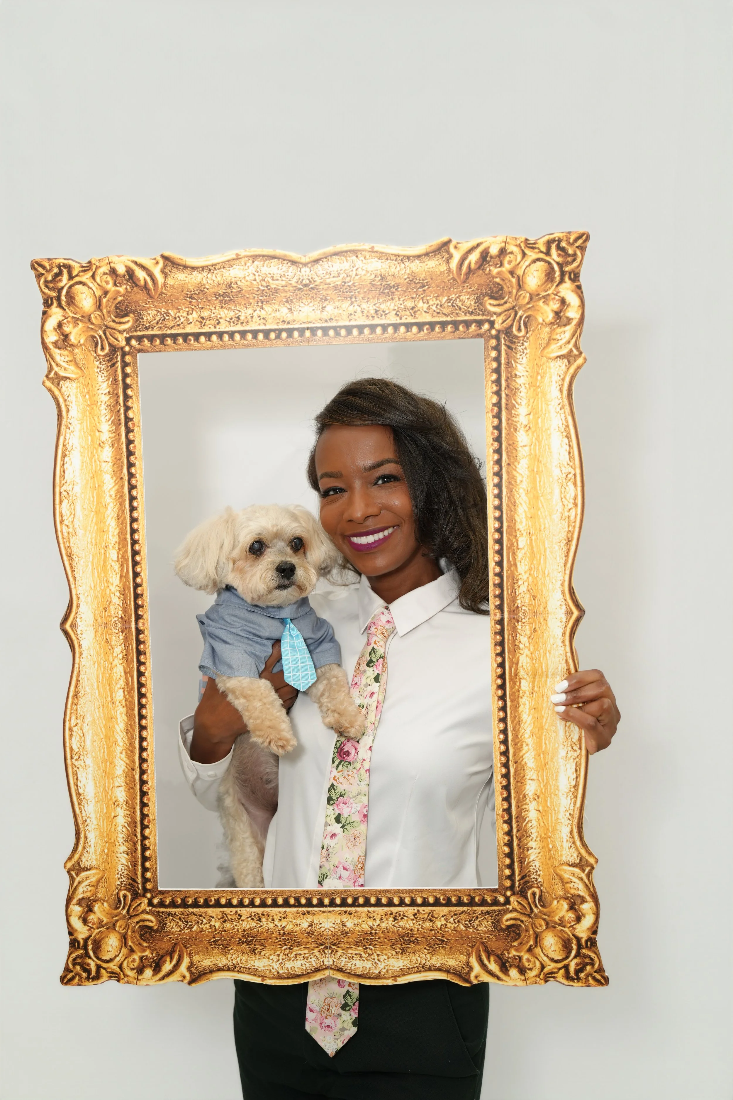 A woman with dark hair, smiling, holding a small dog in a blue shirt and tie, standing behind an ornate gold frame.