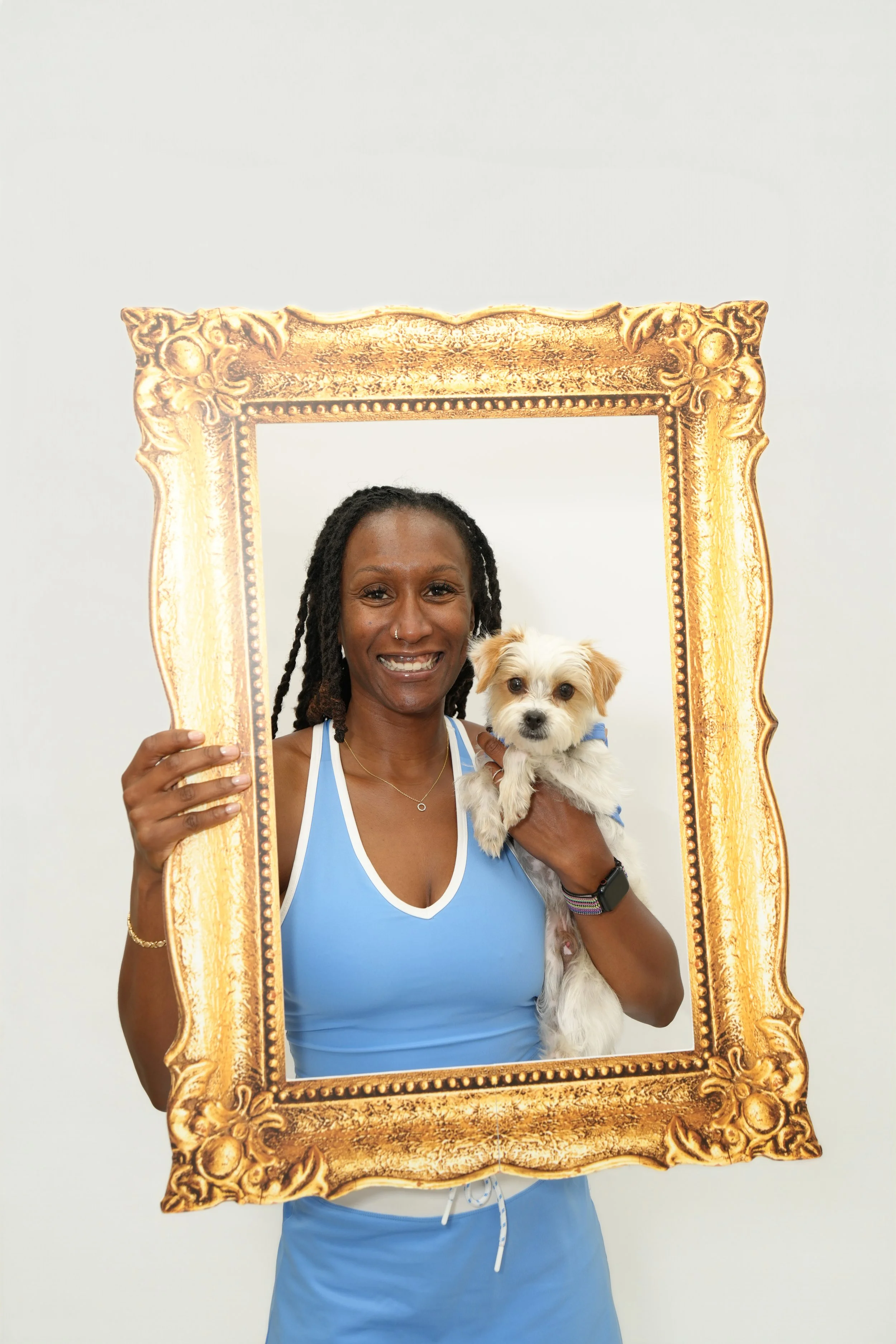 A woman in a blue sports bra and blue shorts holding a small white dog, posing behind a large ornate gold picture frame against a plain white wall.